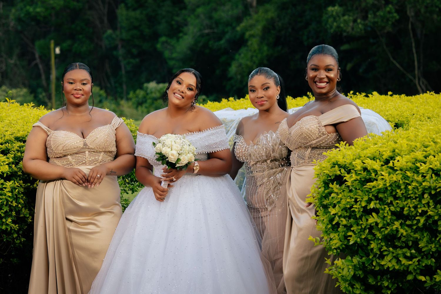 Bride in white gown poses with three bridesmaids in champagne dresses outdoors among lush greenery.