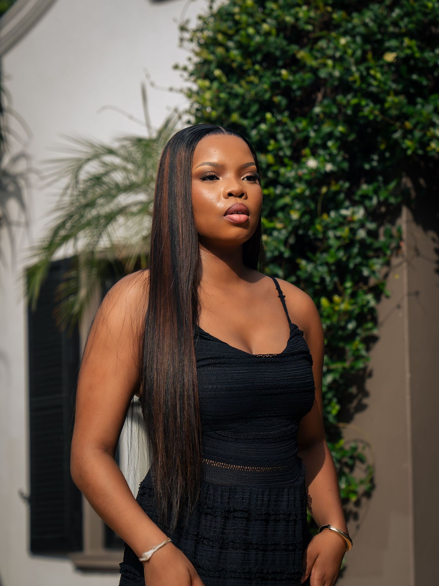 Young Black woman in black dress with long straight hair poses outdoors near white building and green bushes.