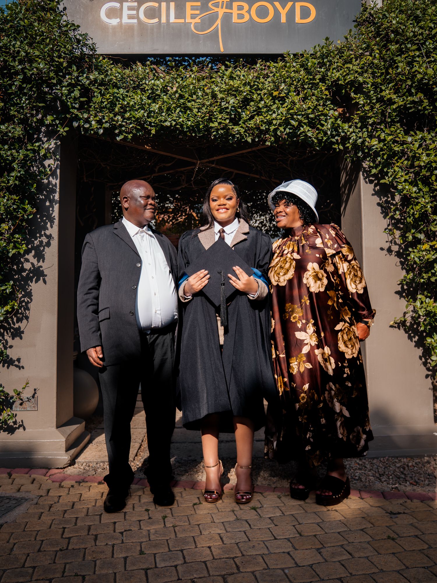 Graduate in cap and gown poses with two proud family members outside Cecile & Boyd venue.