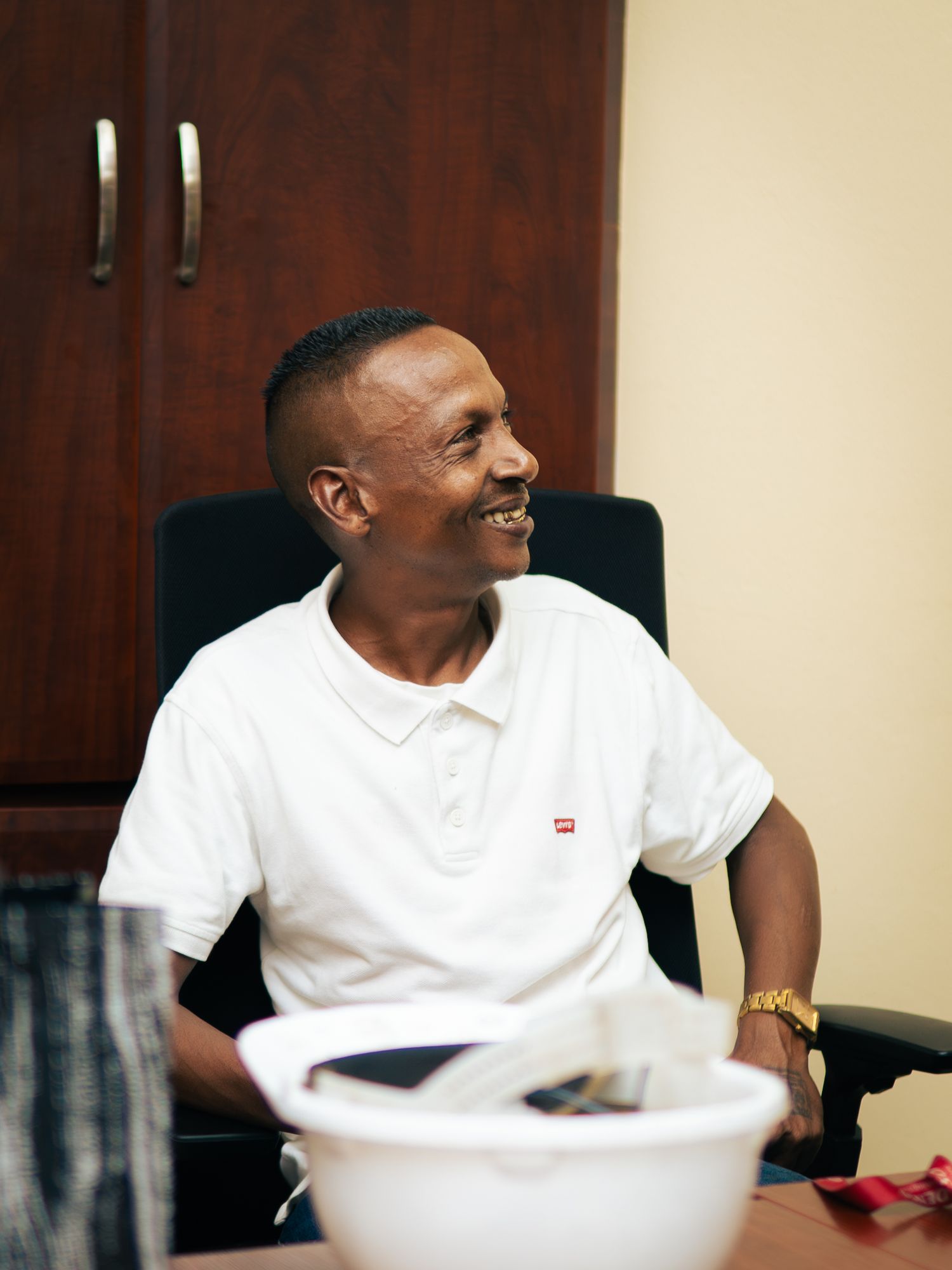 A smiling man in a white Levi's polo shirt sits relaxed in a black office chair near a wooden cabinet.