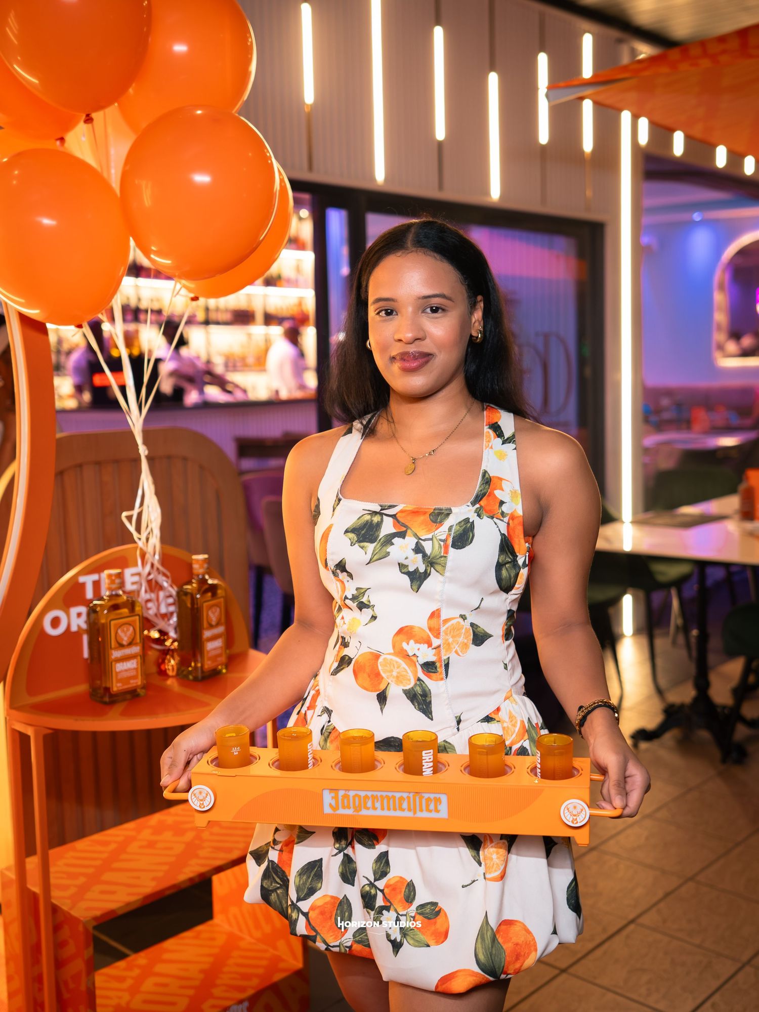 Woman in orange floral dress holding Jagermeister shot tray surrounded by orange balloons at a branded event.