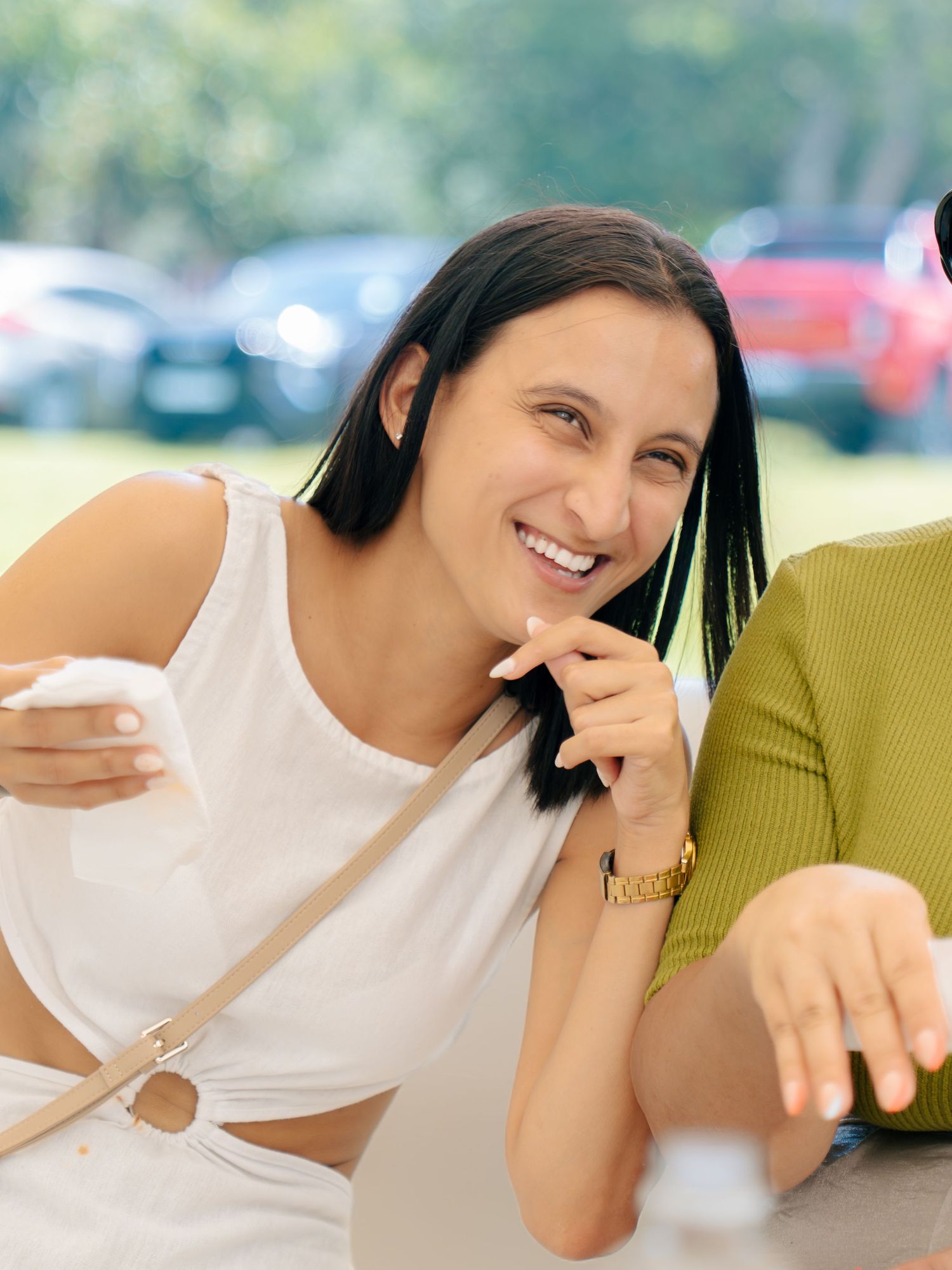 Smiling woman in white crop top with crossbody bag sitting outdoors near parked cars on a sunny day.