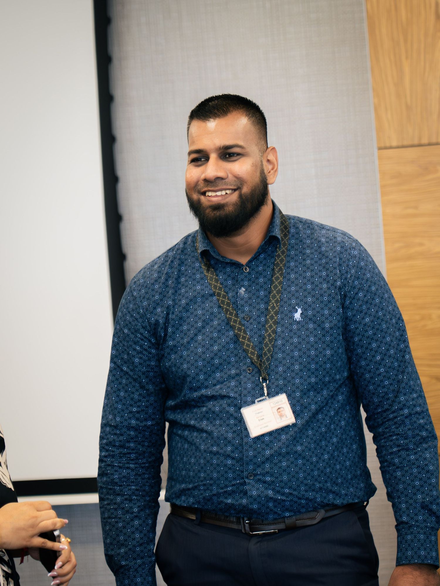Smiling man in blue patterned shirt wearing a lanyard badge stands in a modern conference room.