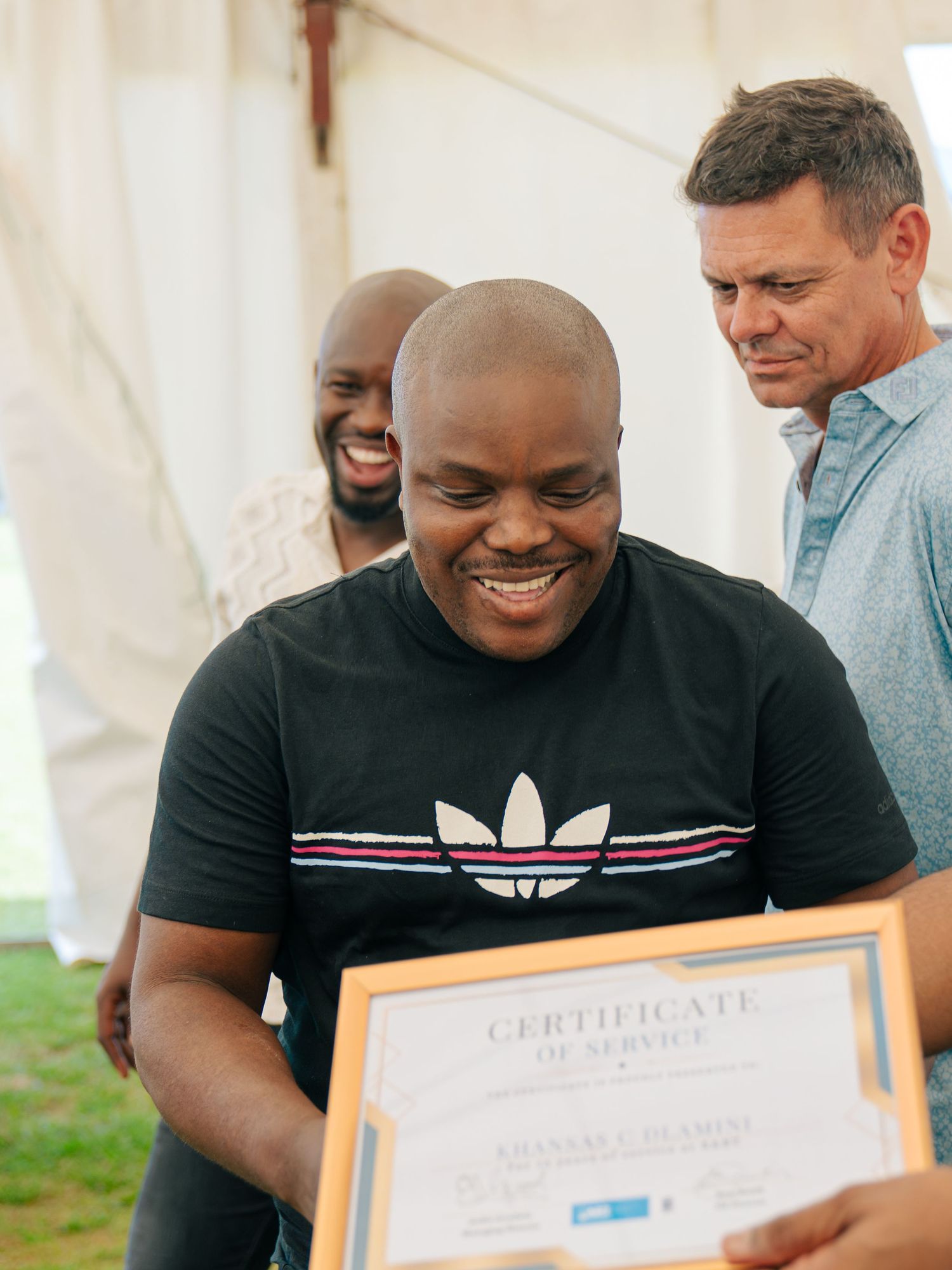 Smiling man in Adidas shirt proudly holds a Certificate of Service award at an outdoor ceremony.