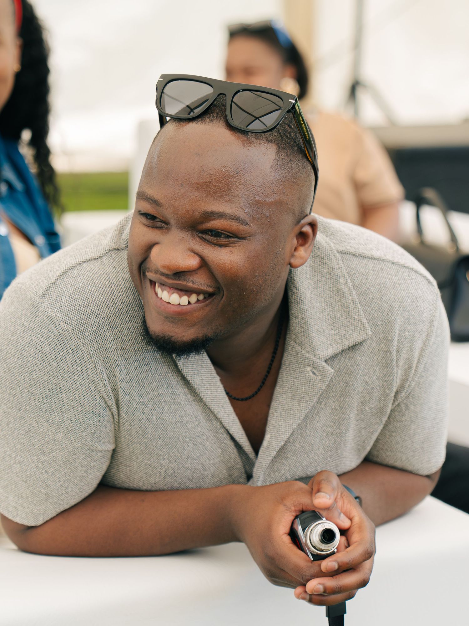 Smiling young Black man with sunglasses on head holds camera, wearing gray shirt at outdoor event.