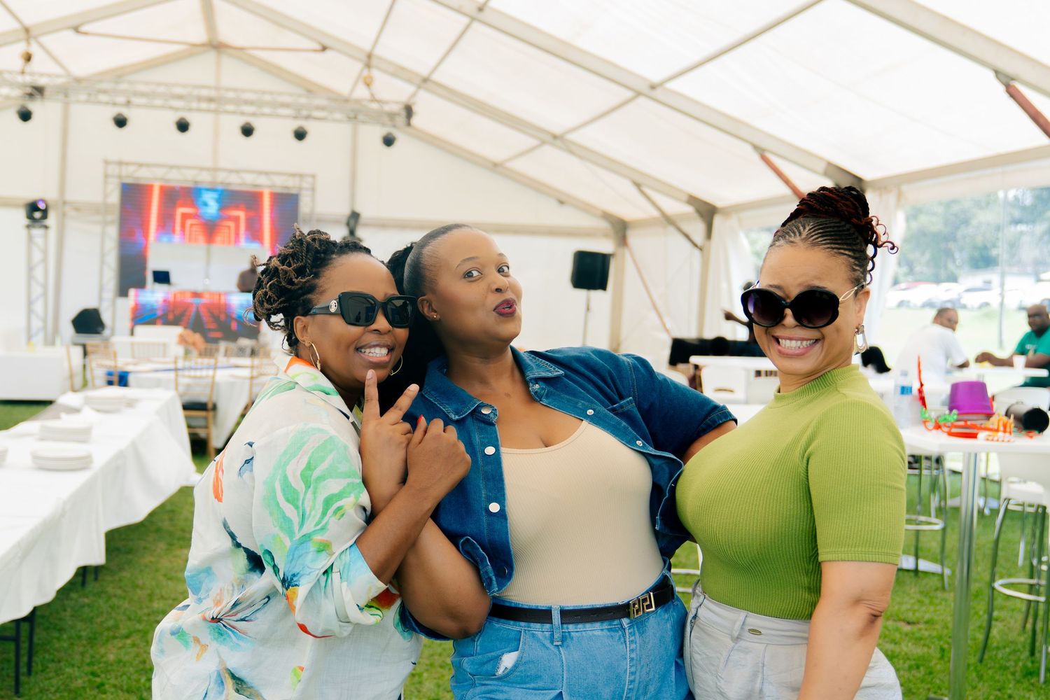 Three women posing together at an outdoor festival event under a white tent, wearing stylish summer outfits and sunglasses.
