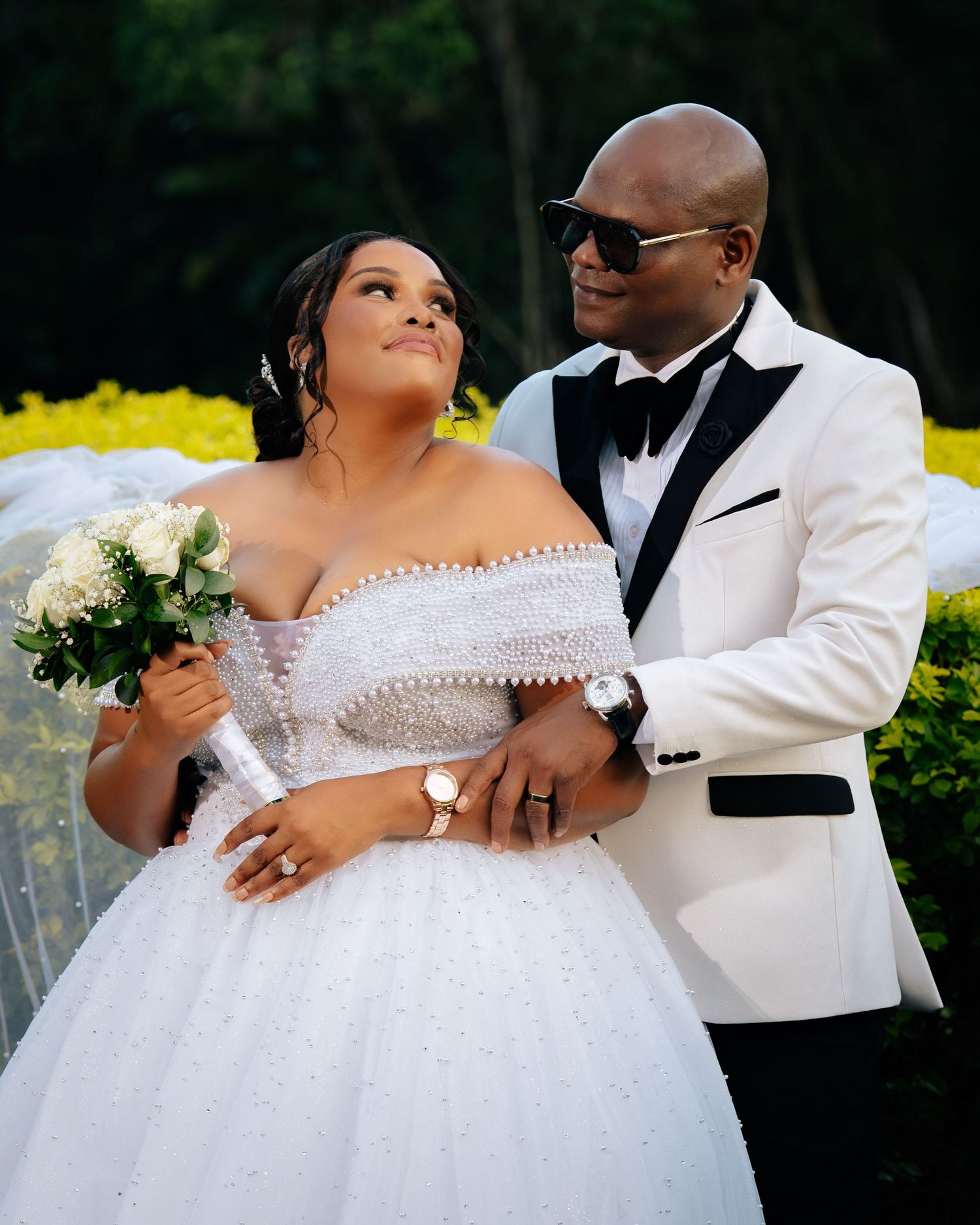 Elegant wedding couple in white attire, bride holding bouquet, groom in tuxedo, sharing a romantic glance outdoors.