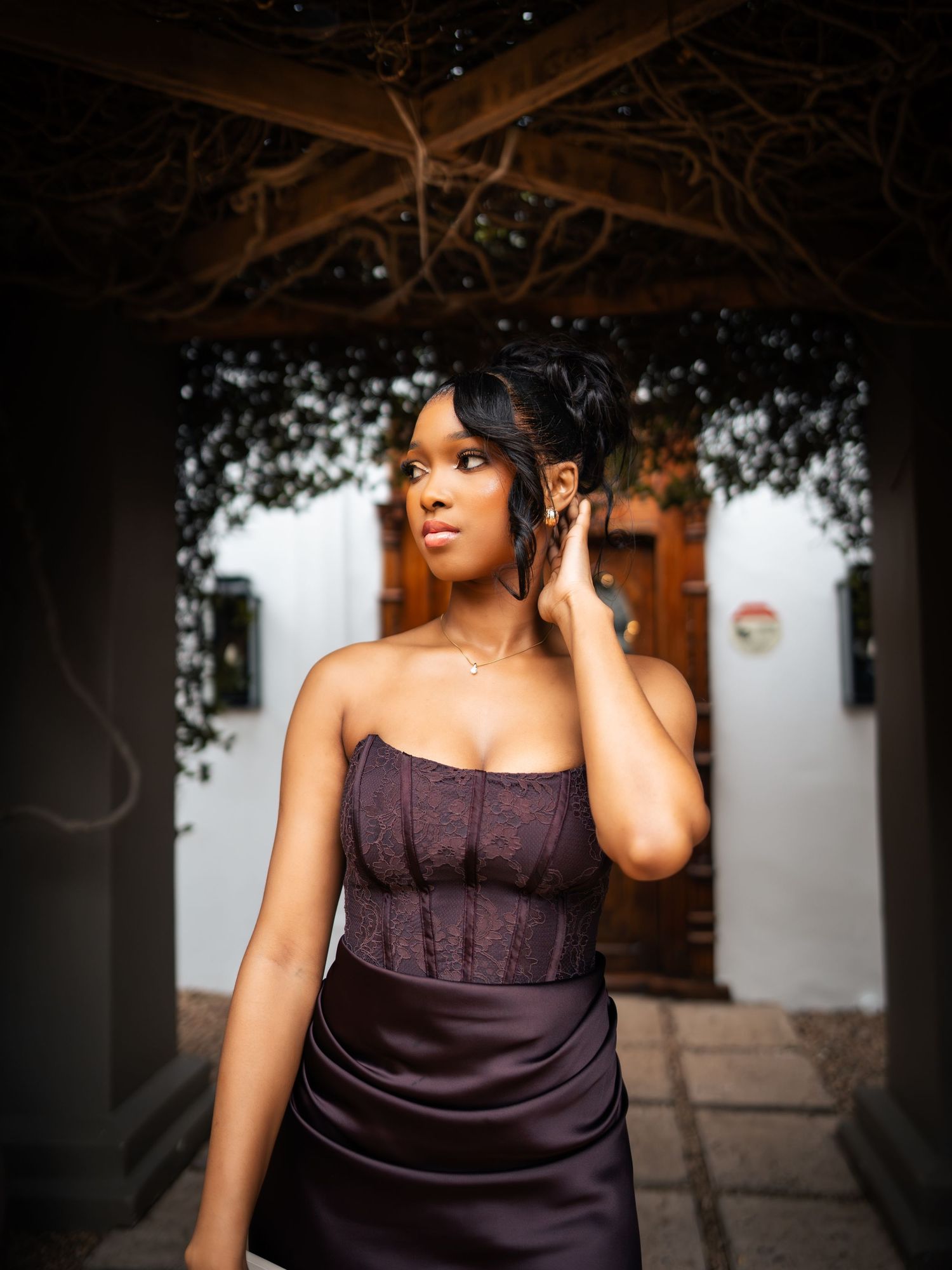 A young Black woman in a strapless purple corset and satin skirt poses elegantly outdoors near a rustic archway.