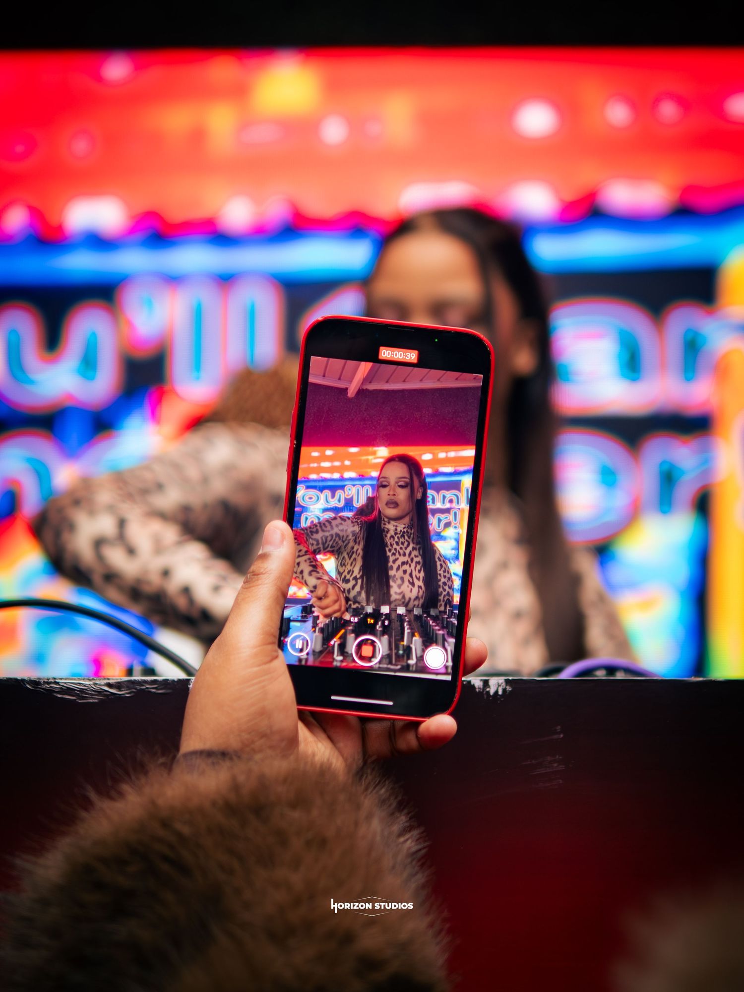 A hand holds a smartphone capturing a woman DJ performing on stage at a colorful neon-lit event.