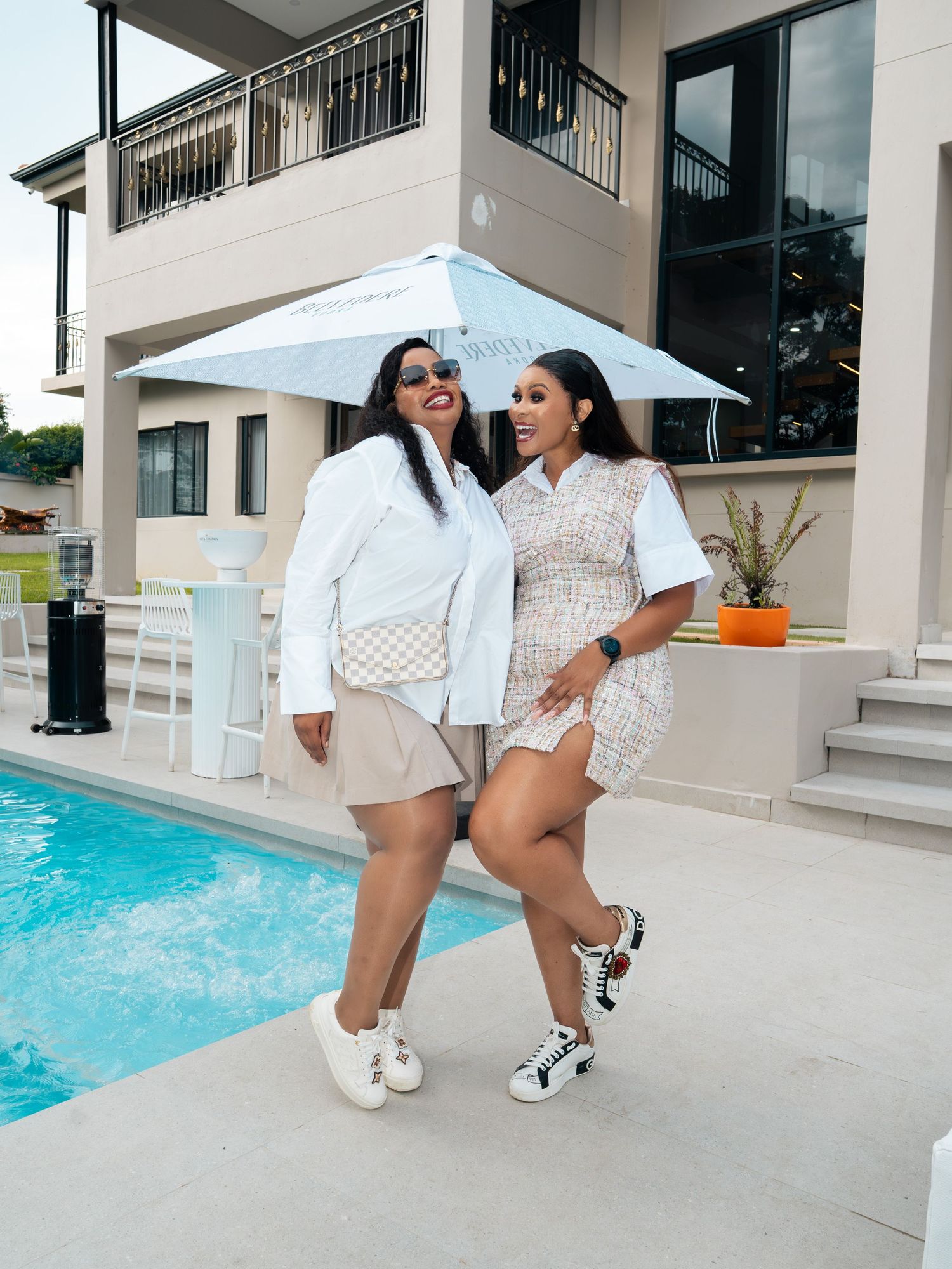 Two stylish women posing by a luxury pool, wearing white outfits and sneakers, with a large umbrella and modern home.