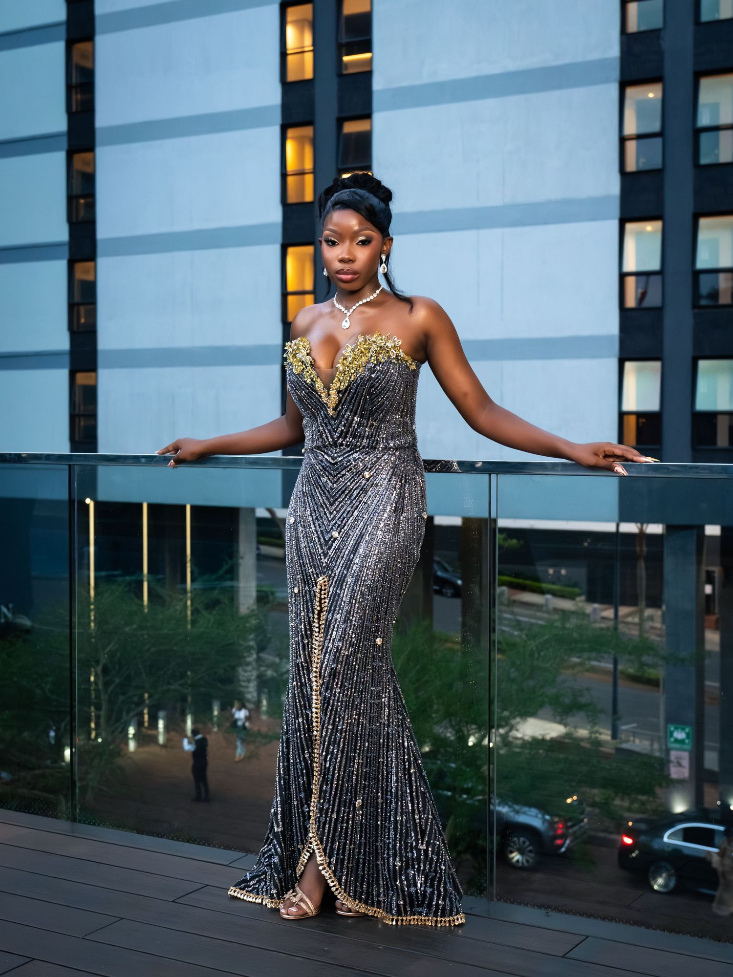 Woman in elegant sequined gown with gold detailing poses on a balcony at dusk.