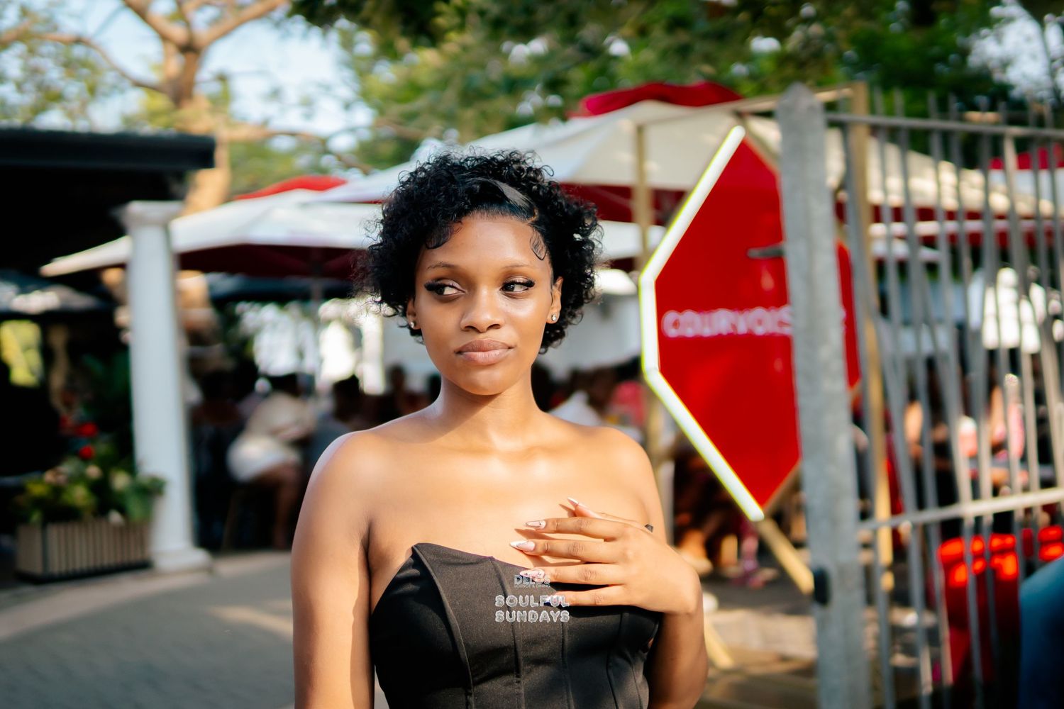 Young Black woman in strapless black dress poses outdoors near a red stop sign at a vibrant street market.