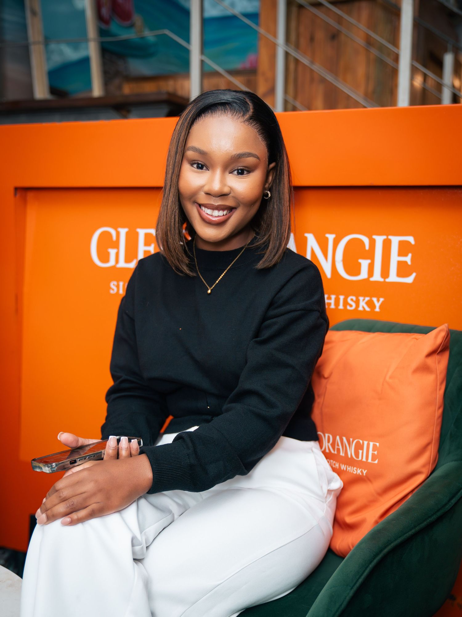 Smiling woman in black top and white pants seated at a Glenmorangie Scotch Whisky branded orange booth.