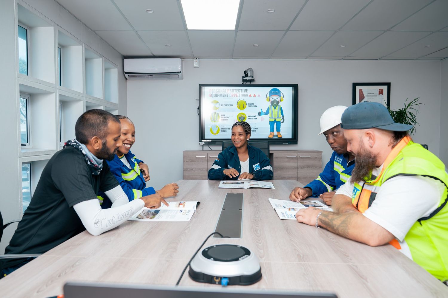 Diverse team in safety vests discussing workplace safety training in a modern conference room with a presentation screen.
