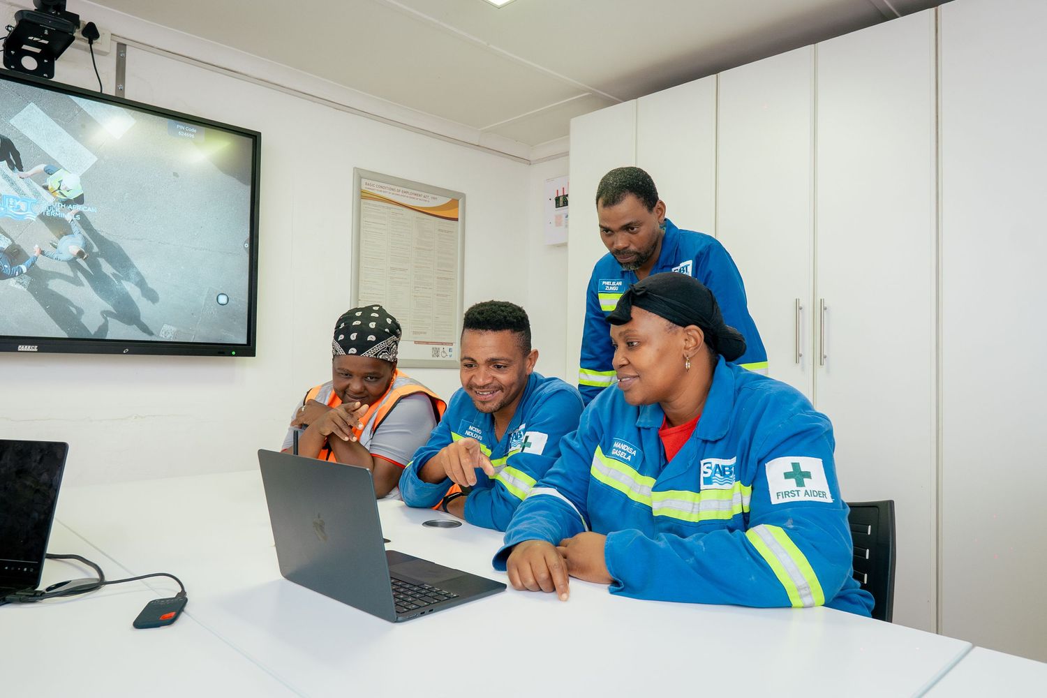 Four workers in blue safety uniforms collaborate around a laptop in a modern office with a monitor on the wall.