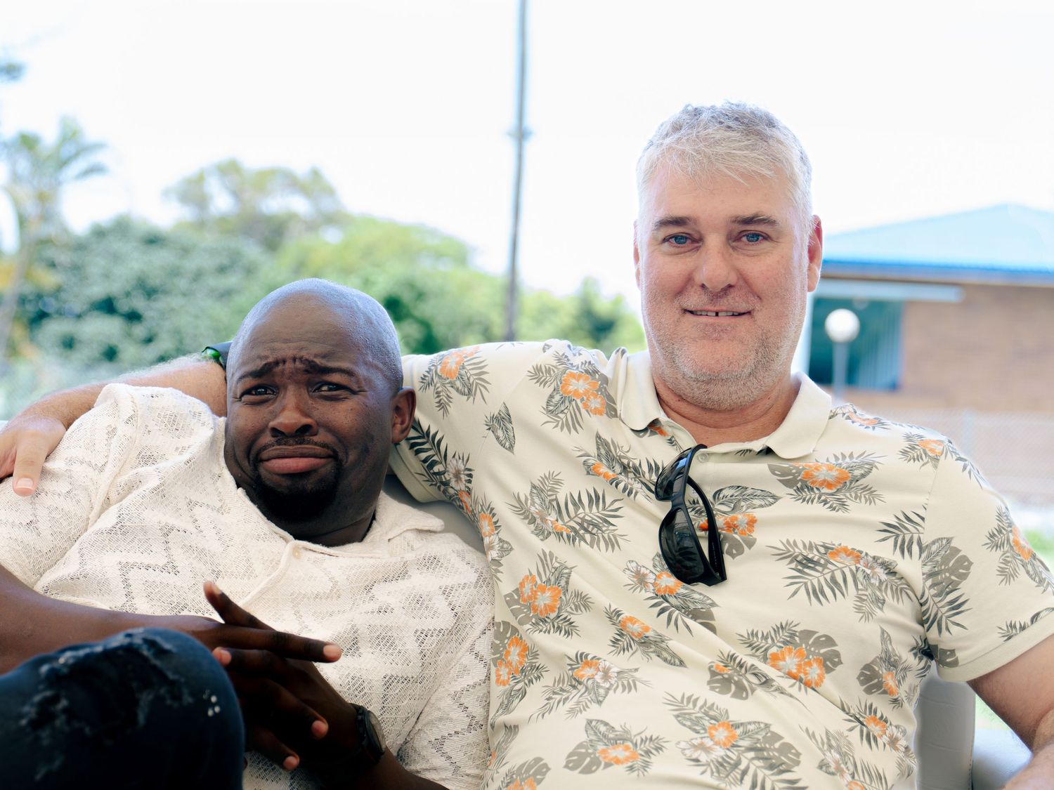 Two smiling men pose together outdoors, one wearing a floral Hawaiian shirt, with a blue building and trees in the background.