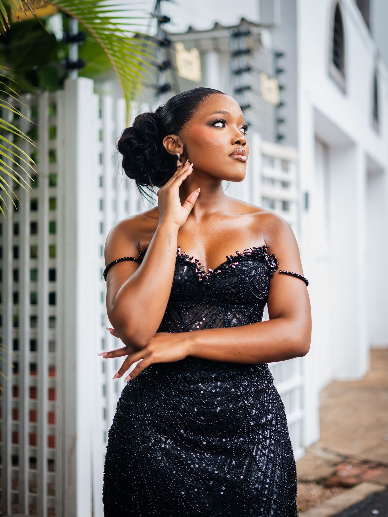 Elegant woman in black sequin off-shoulder gown posing gracefully outdoors near white lattice wall.