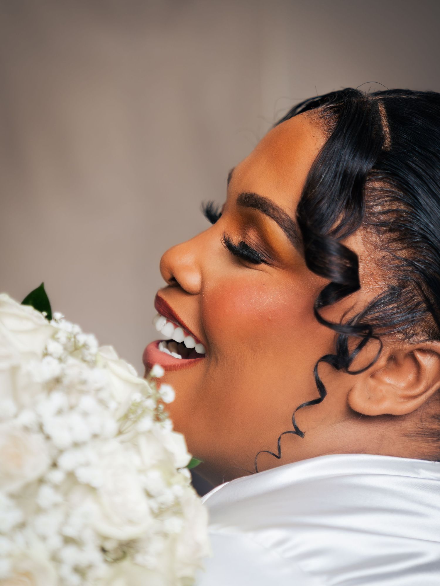 A joyful Black bride in profile laughing with elegant finger waves, bold lashes, and holding a white floral bouquet.