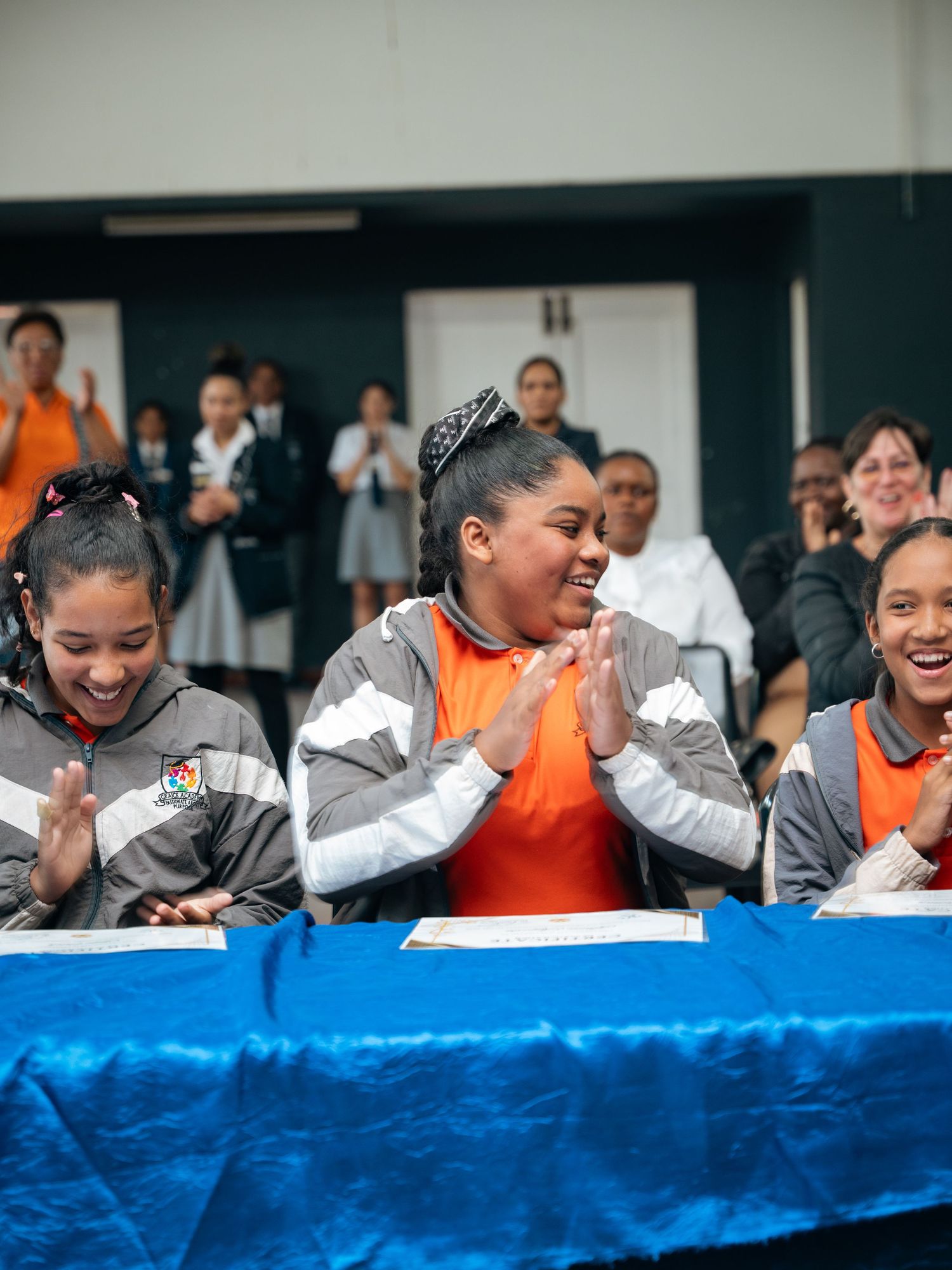 Three young women in matching gray jackets laugh and clap at a blue-draped table during a celebratory signing event.