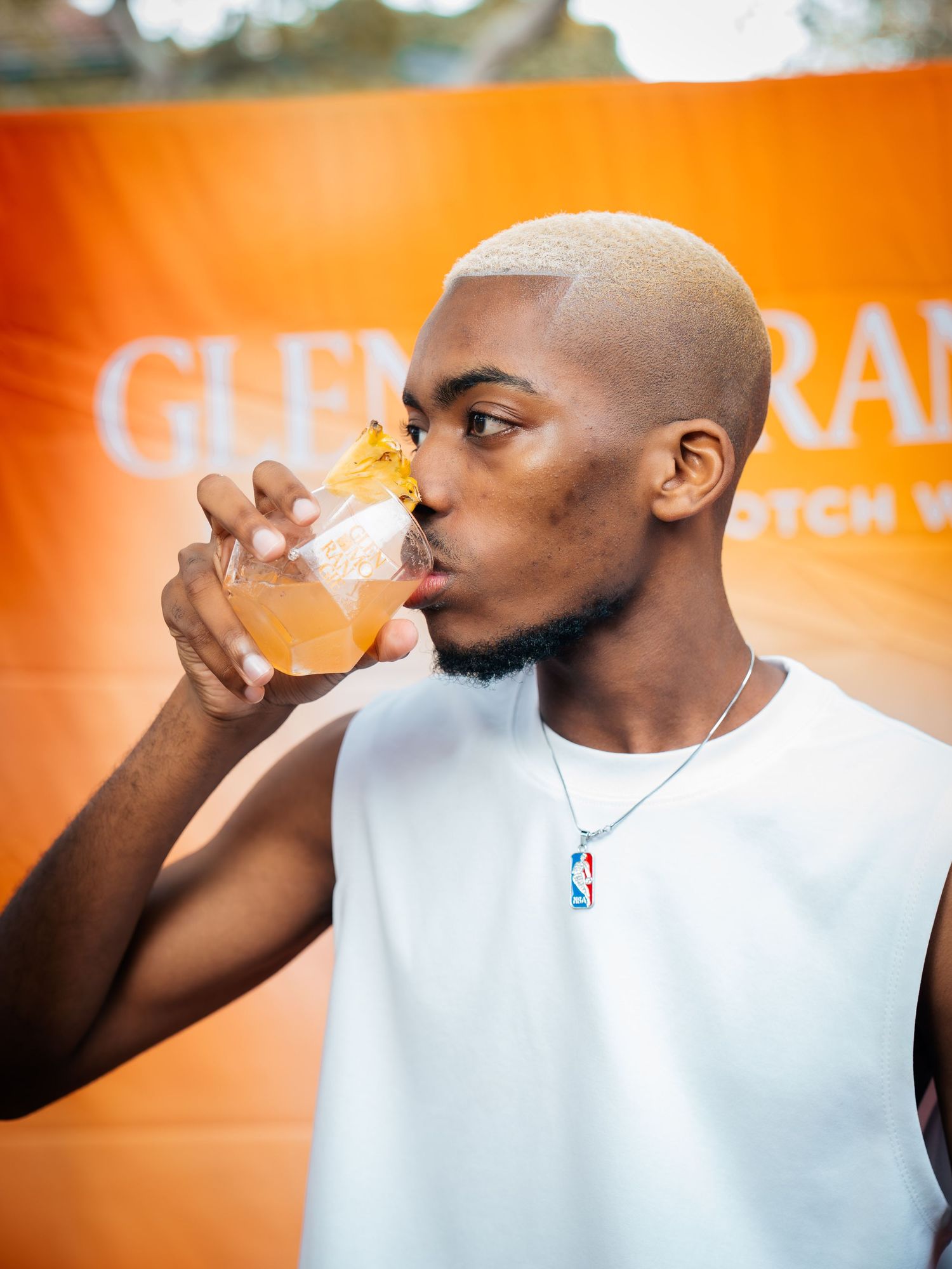 Young man drinking an orange cocktail at a Glenfarclas Scotch whisky event wearing a white tank top.