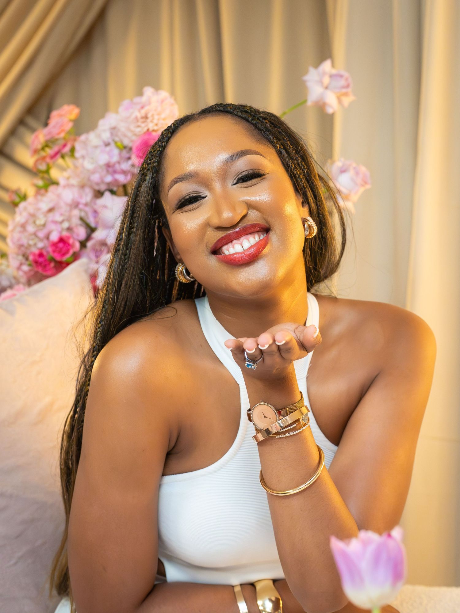 Smiling woman in white top blowing a kiss, wearing gold jewelry, seated near pink floral arrangement.