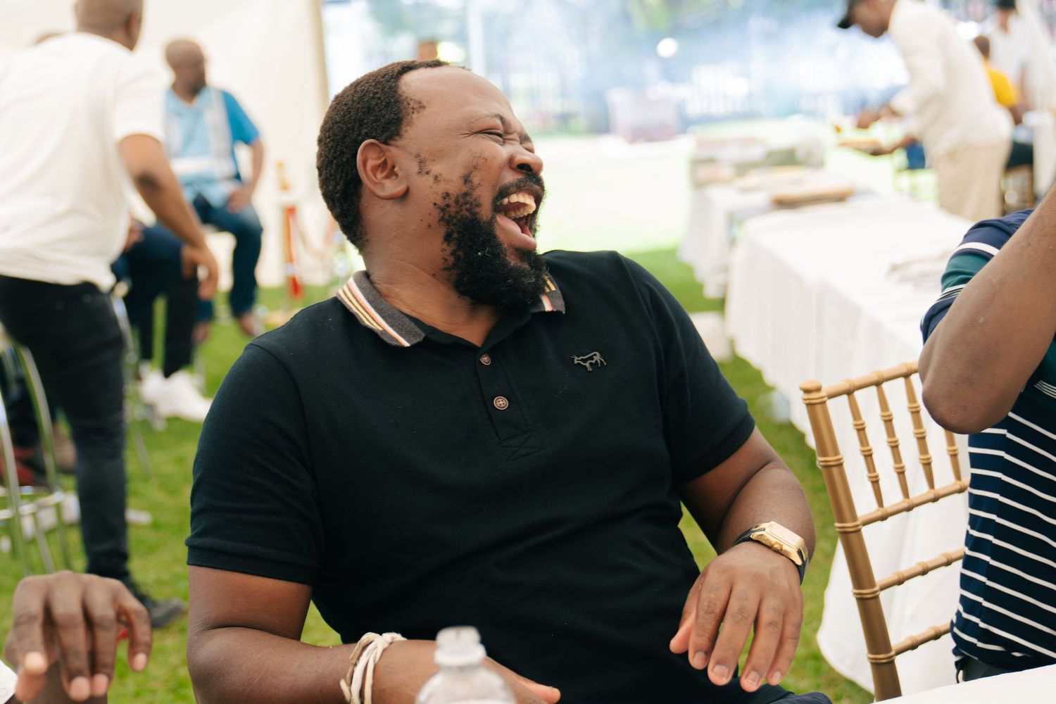 A man in a black polo shirt laughs joyfully at an outdoor event under a white tent.