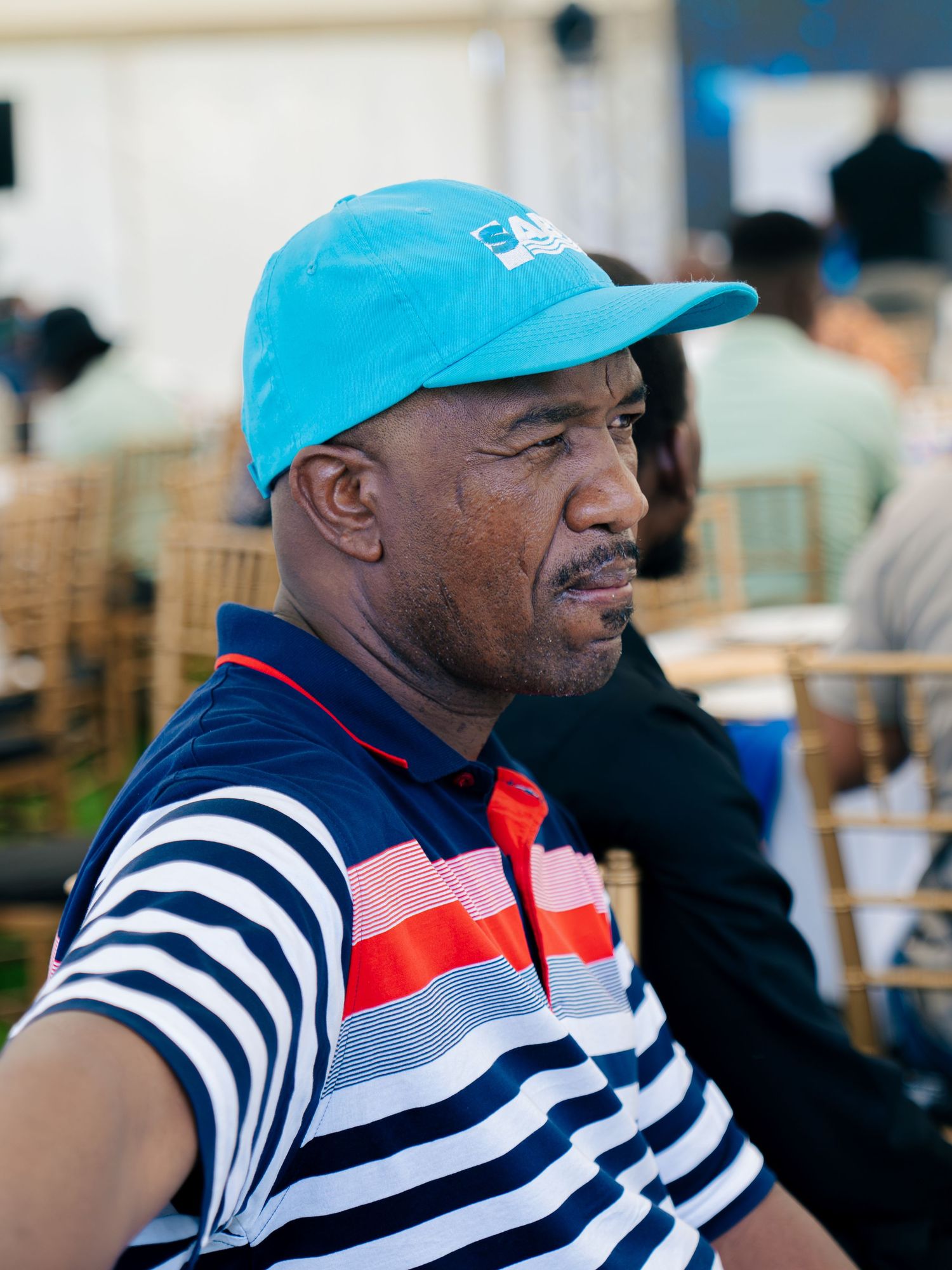 A man wearing a blue cap and striped polo shirt sits at an outdoor event with people in the background.