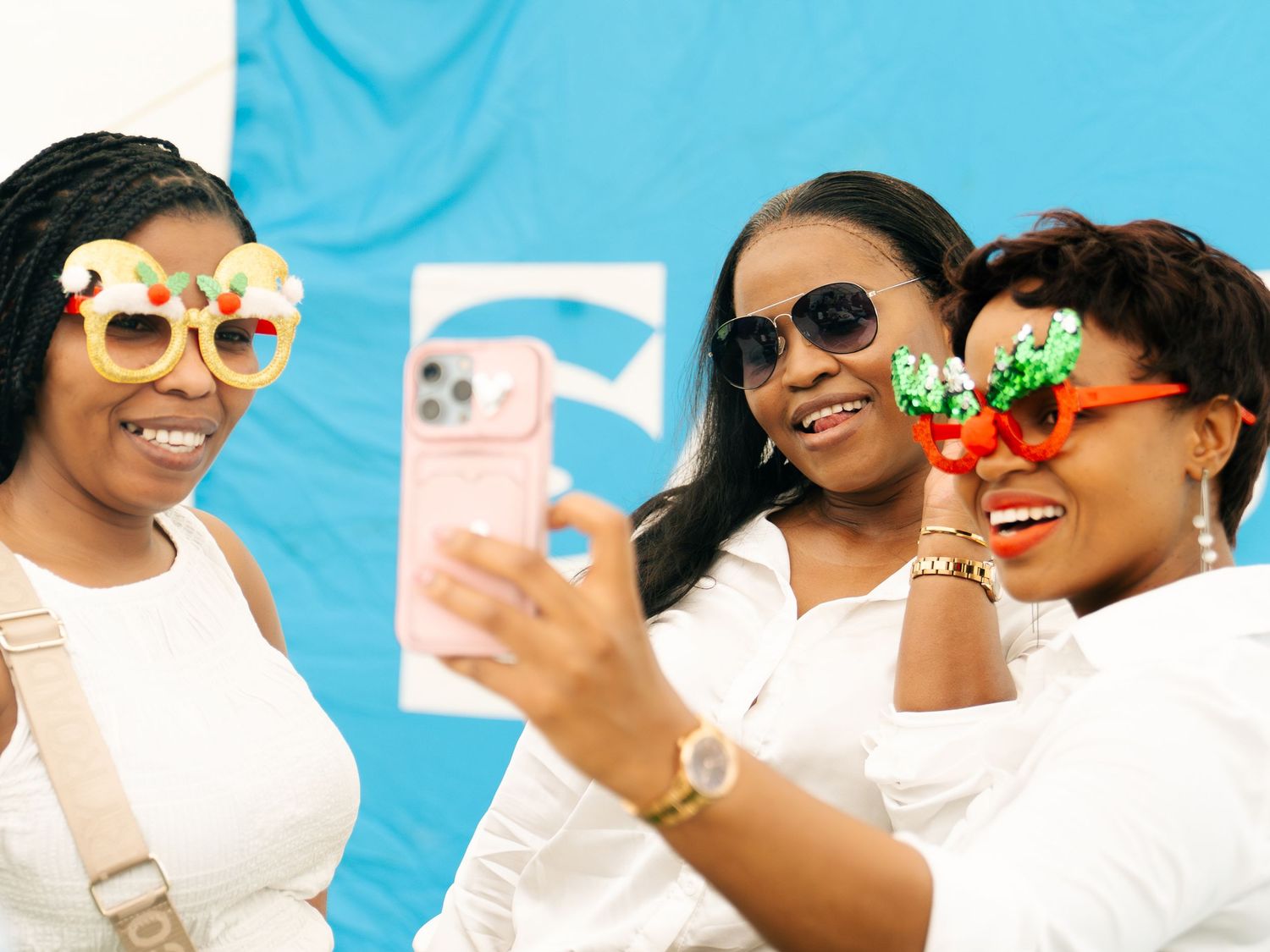 Three women wearing fun novelty sunglasses take a selfie together, smiling and dressed in white outfits.