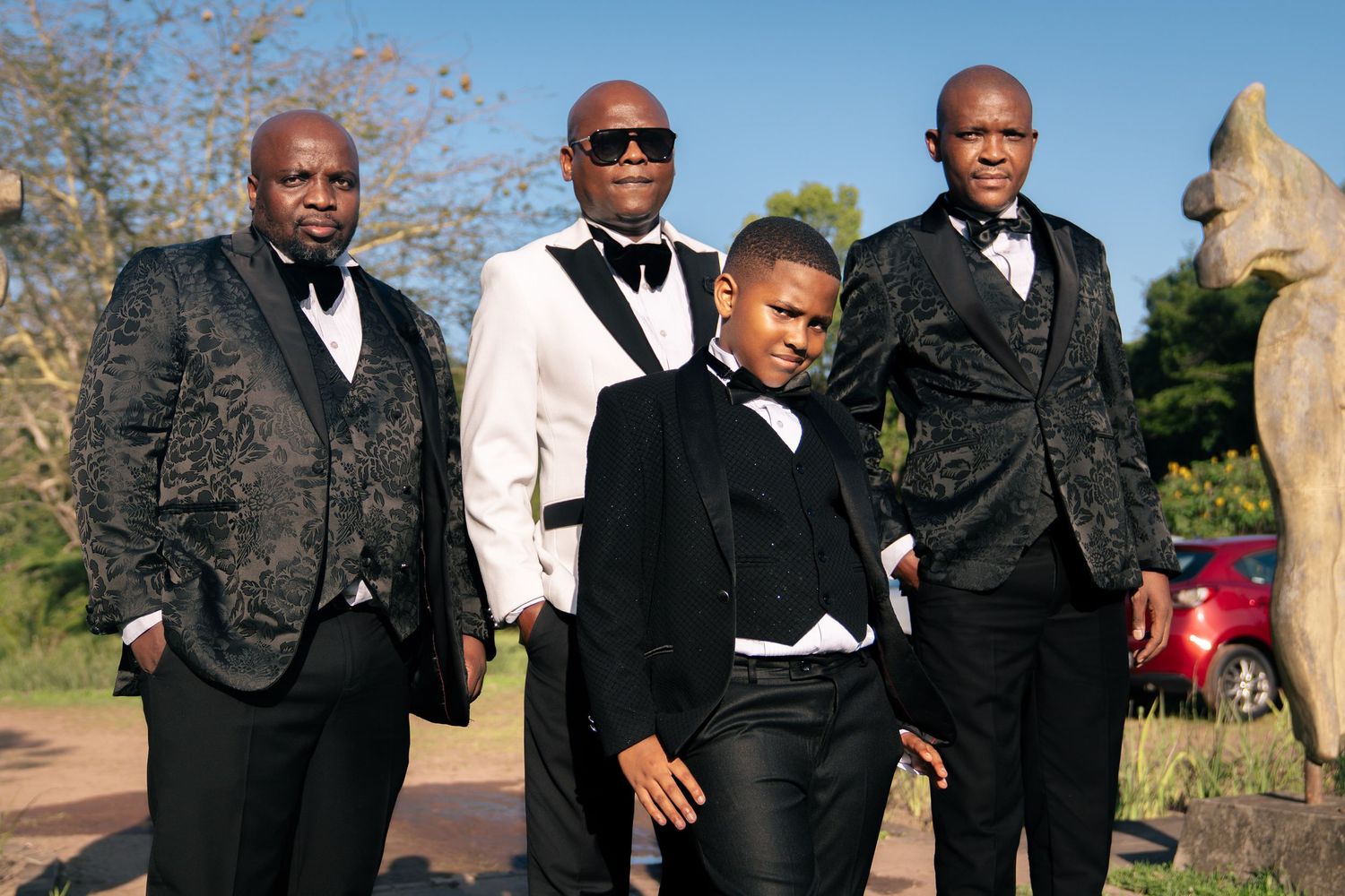 Four well-dressed men in black tuxedos and formal attire posing outdoors for a stylish group portrait.
