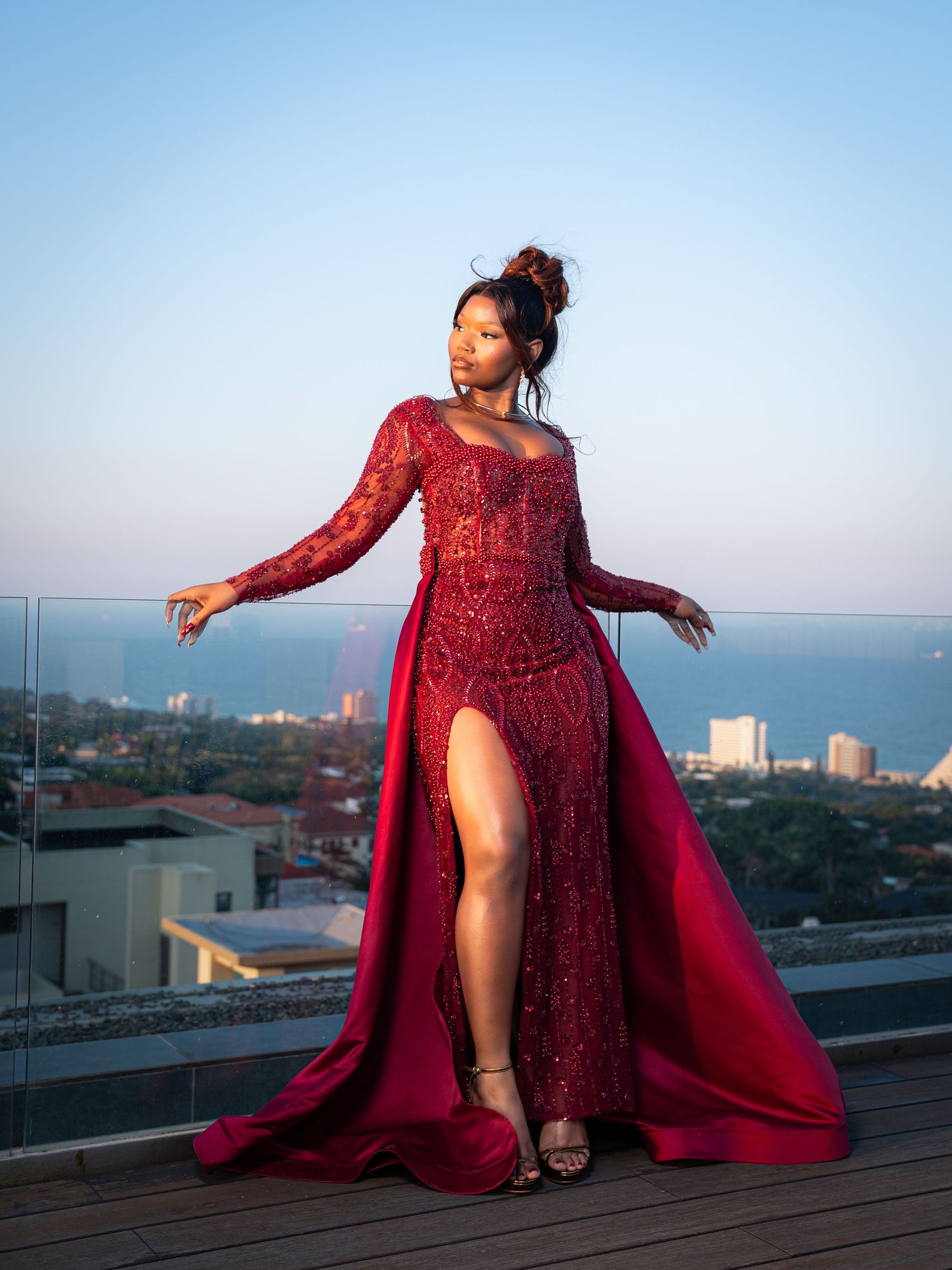 A woman in a stunning red sequined gown with a thigh-high slit poses on a rooftop with a city skyline backdrop.