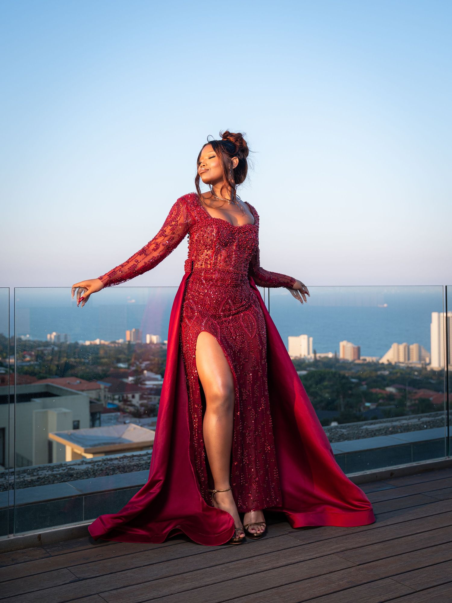Woman in stunning red lace gown with slit posing on rooftop terrace with city and ocean views.