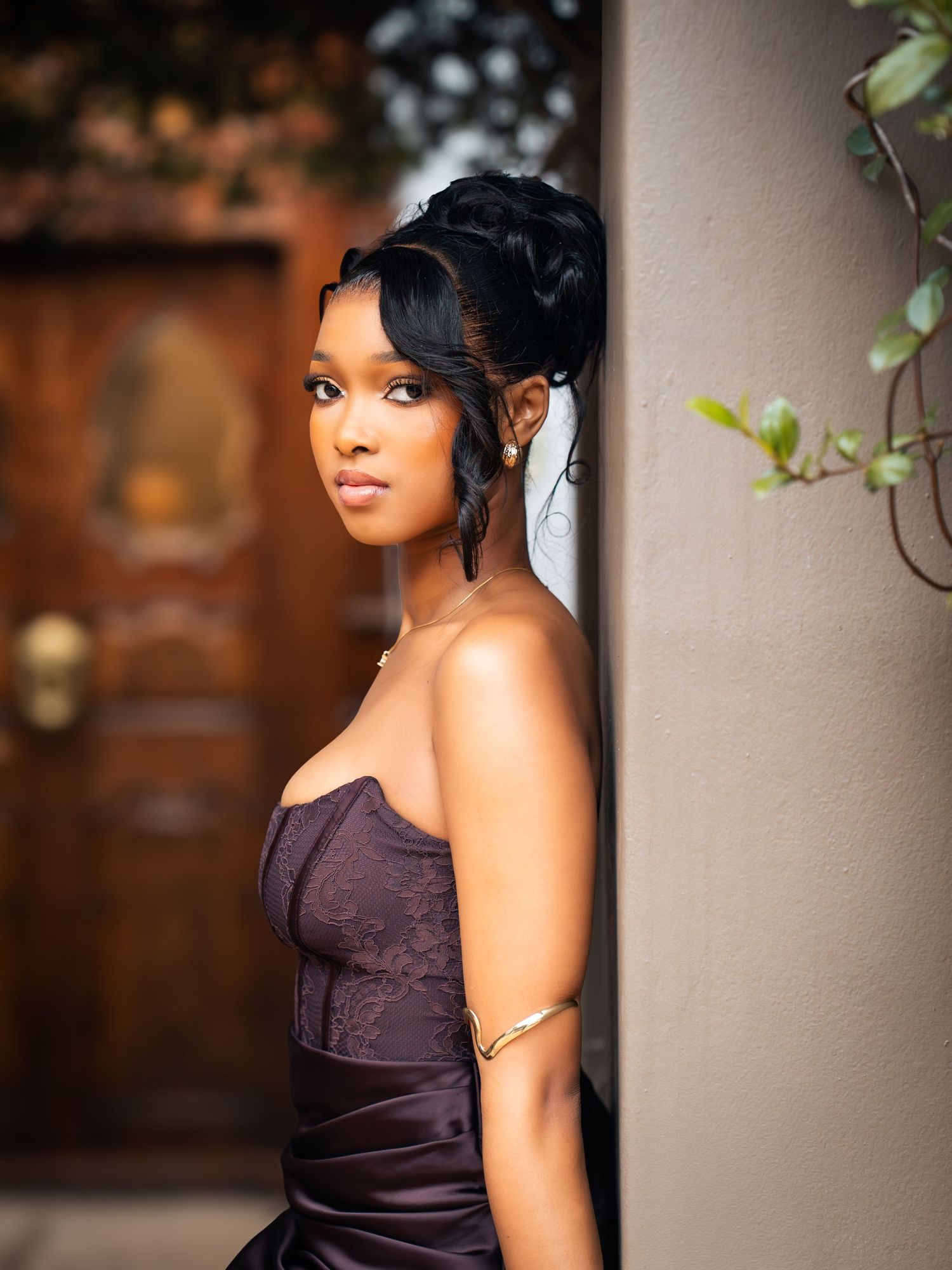 Young woman in strapless purple gown with elegant updo poses gracefully against a wall outdoors.