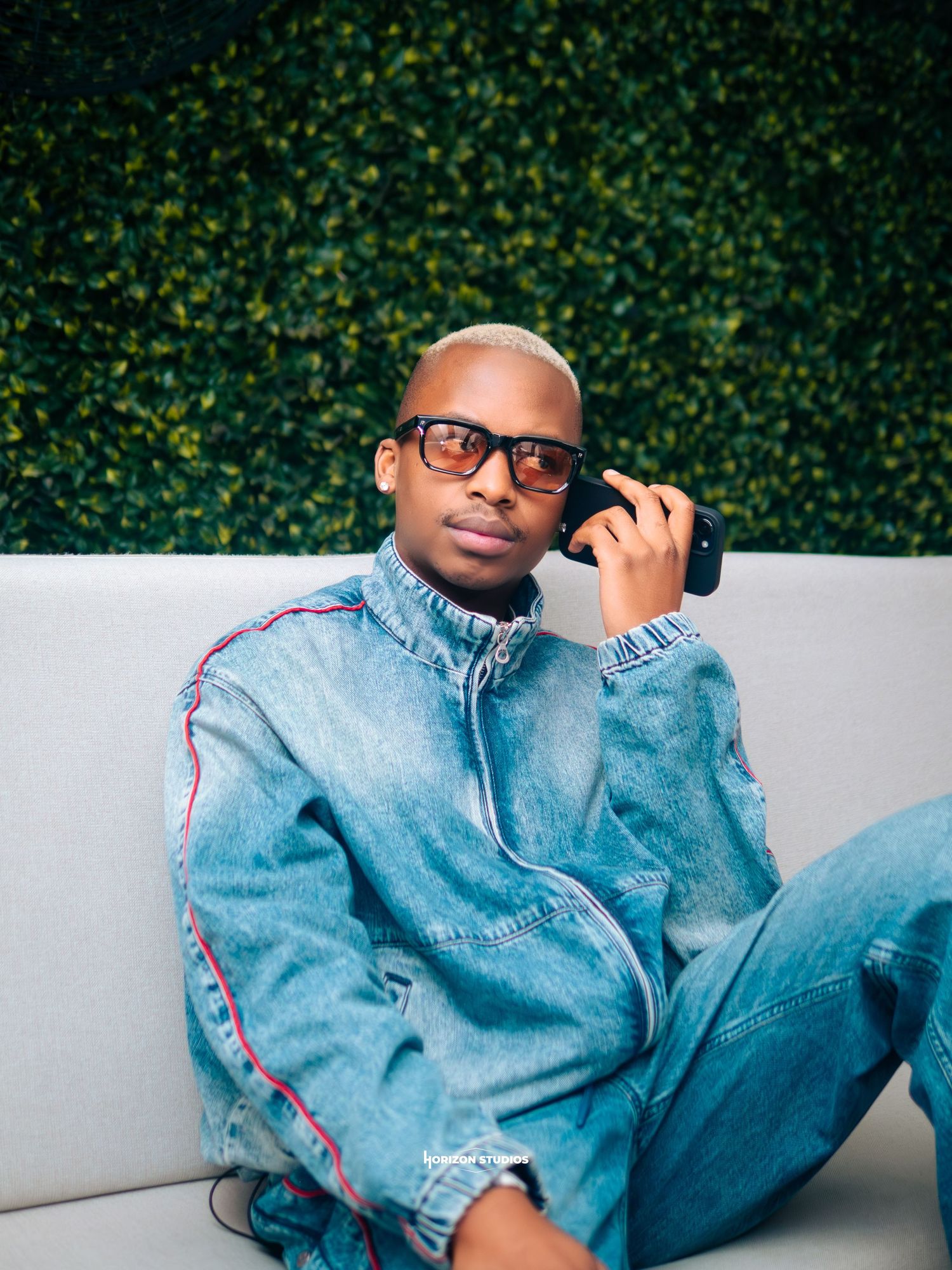 A stylish man in a denim outfit and sunglasses sits relaxed on a white couch against a green hedge wall.