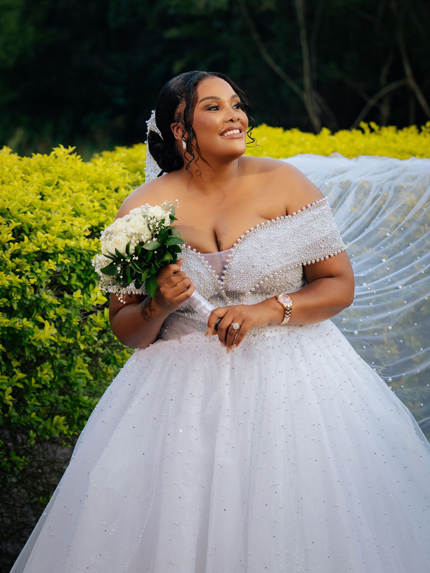 Plus-size bride in a sparkling off-shoulder white ball gown holds a white bouquet, smiling outdoors near yellow hedges.