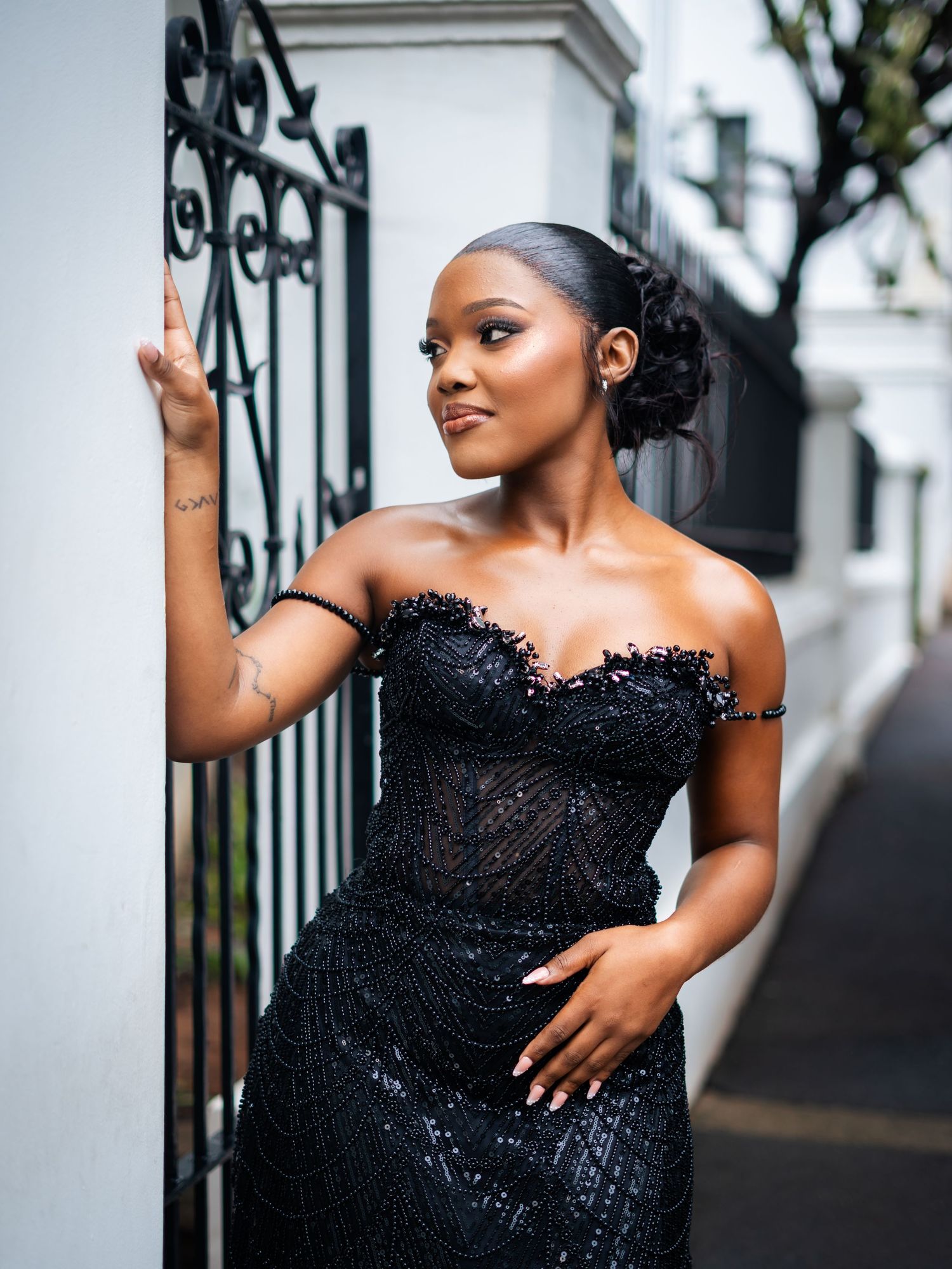 A woman in a black sequined off-shoulder gown poses elegantly by an ornate iron gate.