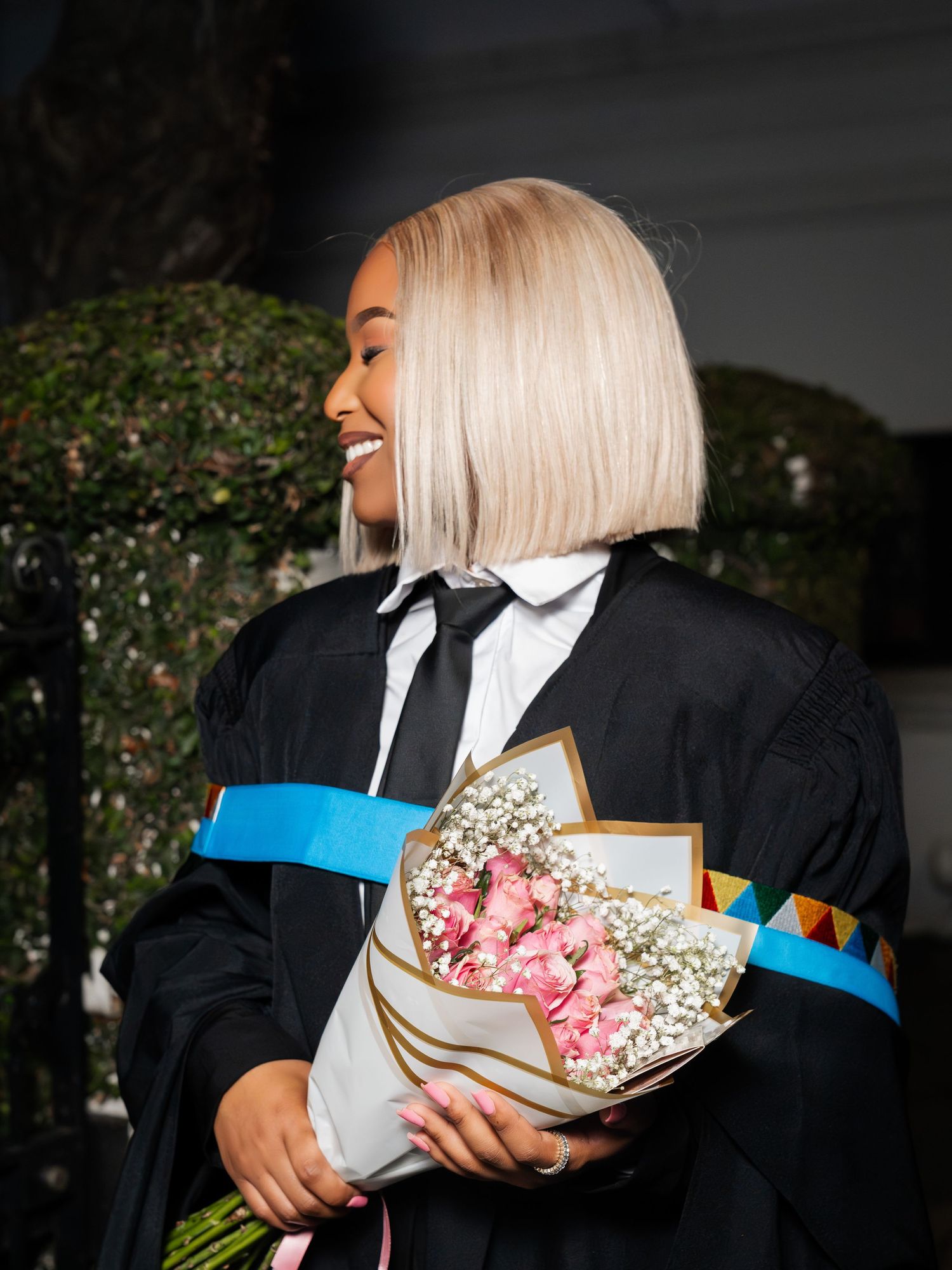 A smiling graduate in black gown with blue sash holds a bouquet of pink roses and baby's breath outdoors.