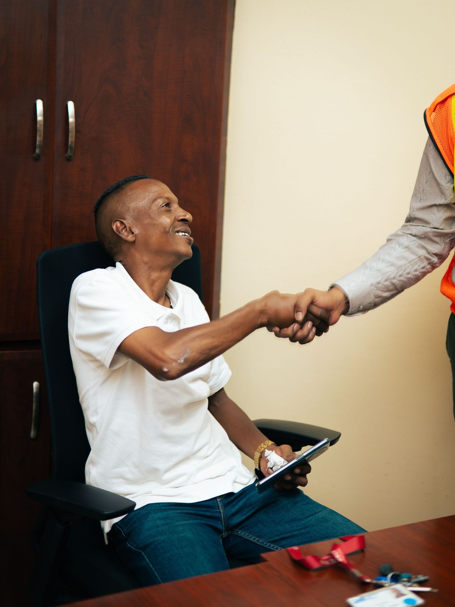 Smiling man in white shirt shakes hands with person in orange vest while seated at office desk holding tablet.