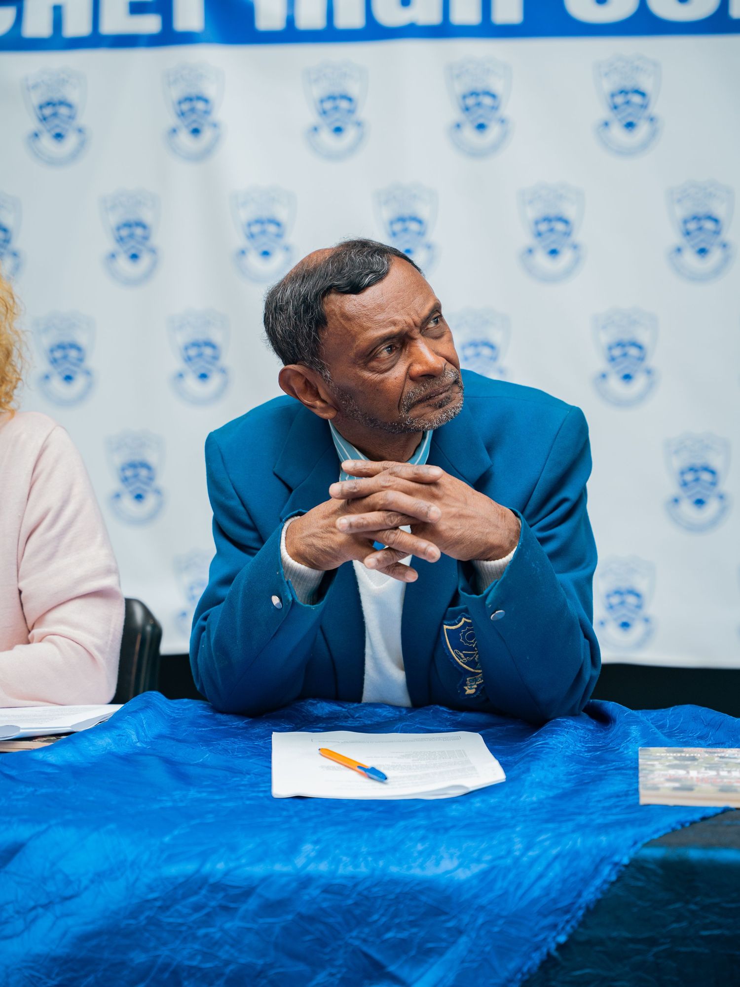 Man in blue blazer sits at a table with papers, hands clasped, in front of a blue branded backdrop.