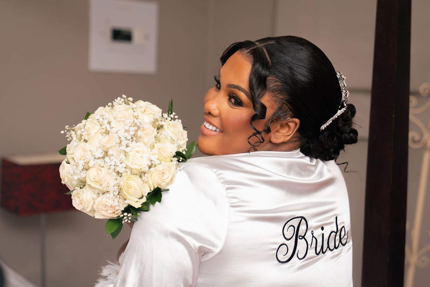 Smiling bride in white satin robe holding elegant bouquet of white roses and baby's breath on her wedding day.