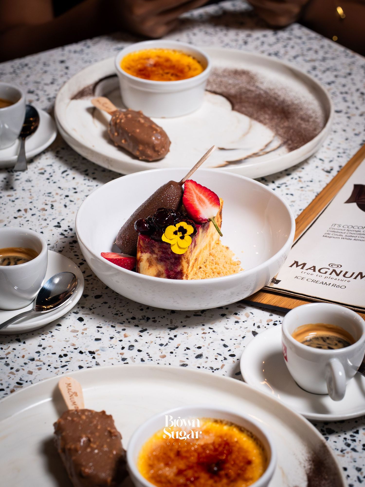 Elegant dessert spread featuring crème brûlée, cheesecake with berries, Magnum ice cream bars, and espresso shots on a terrazzo table.