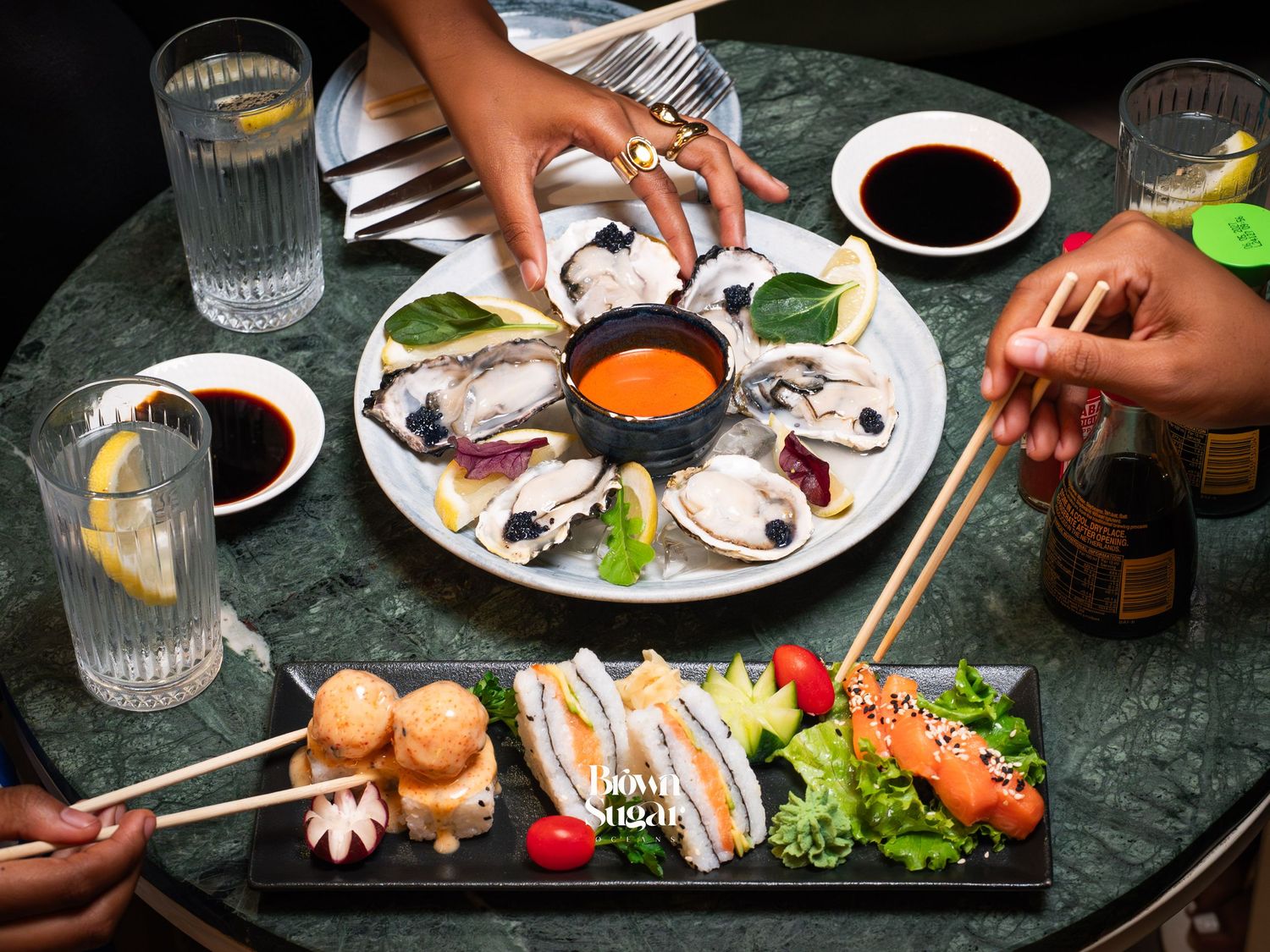 Fresh oyster platter with dipping sauces and sushi rolls served on a marble table with chopsticks and soy sauce.