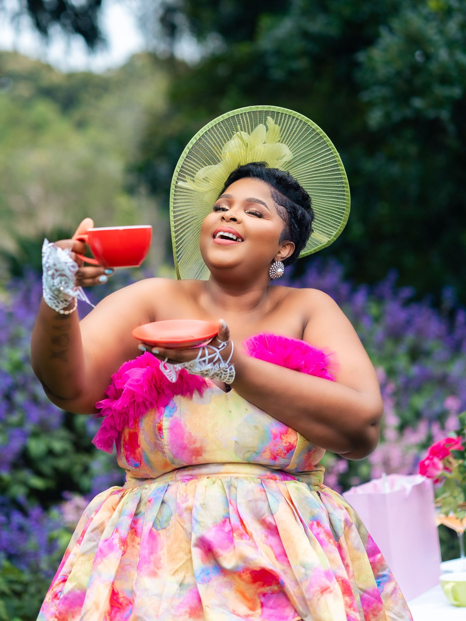 Woman in colorful floral dress and green hat holds red teacup outdoors at elegant garden tea party.