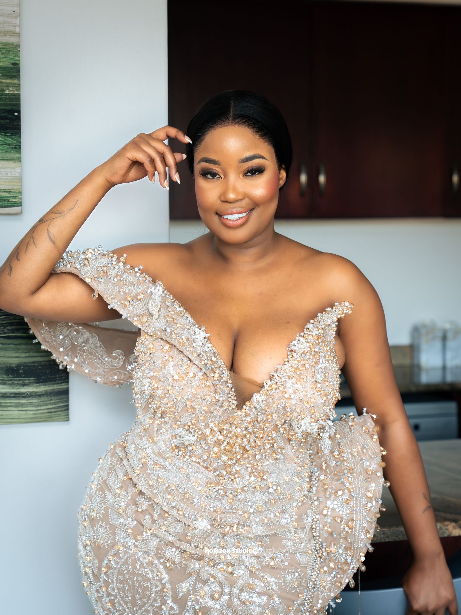 A woman in a beaded off-shoulder gown smiles elegantly, showcasing glamorous bridal fashion and flawless makeup.