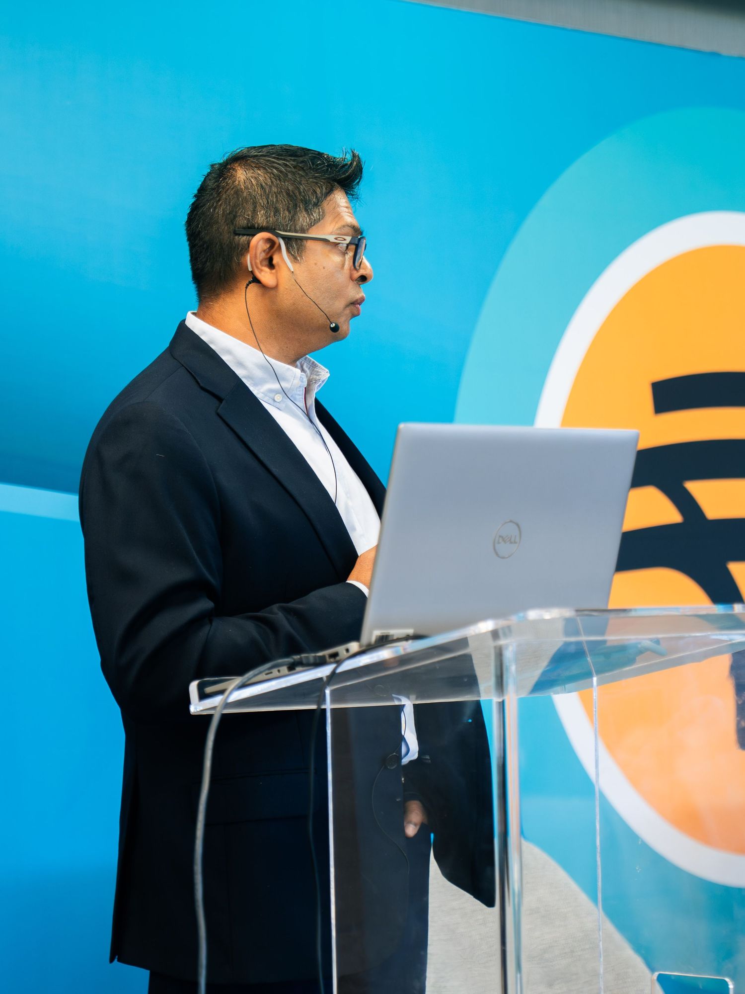 Professional speaker presenting at podium with laptop, wearing black blazer and headset mic against blue backdrop.