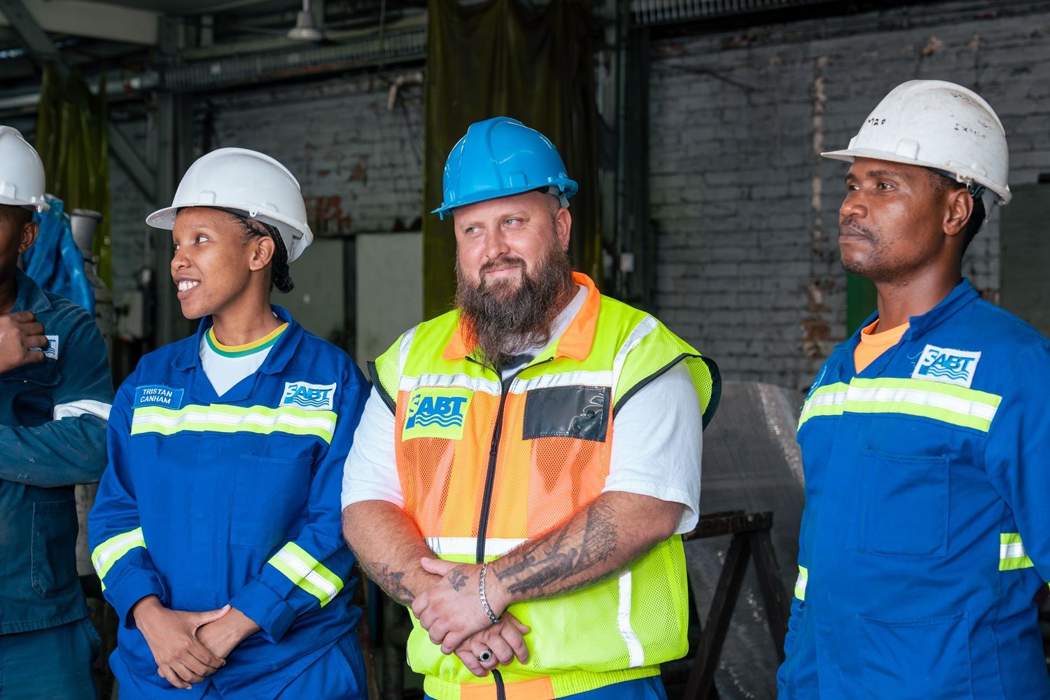 Three workers in hard hats and safety gear pose together at an industrial facility.