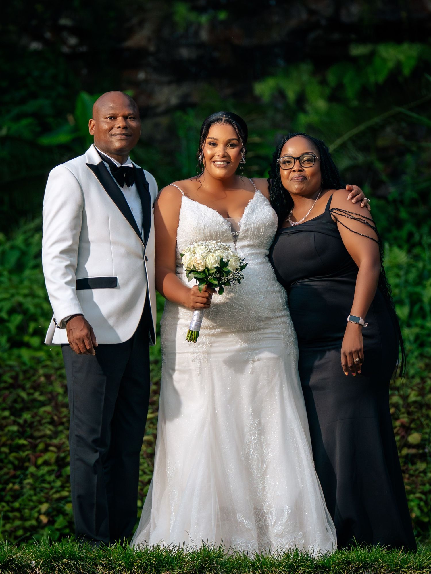 Bride in white gown holds bouquet flanked by man in tuxedo and woman in black dress at outdoor wedding.
