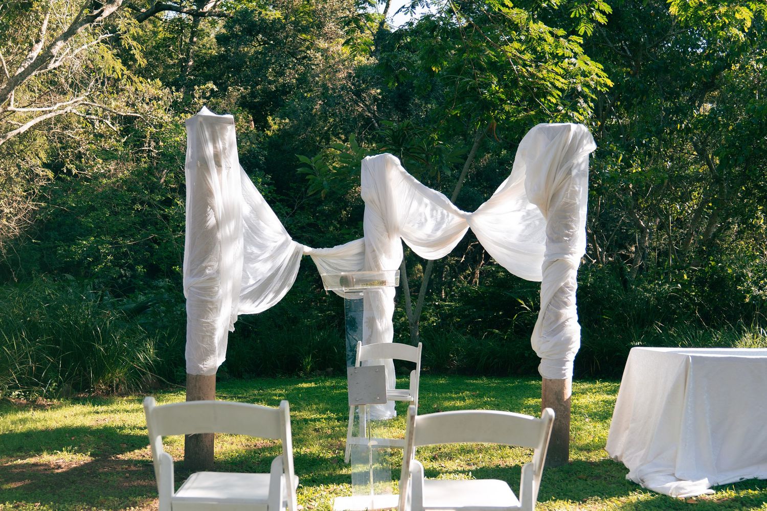 Outdoor wedding ceremony arch draped with white fabric on green lawn surrounded by lush trees.