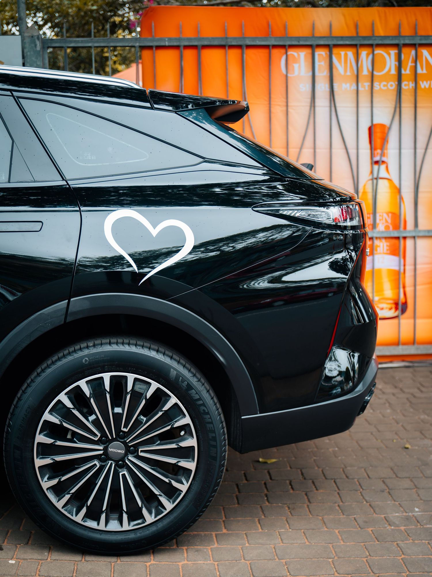 Black luxury SUV with heart decal parked near Glenmorangie whisky advertisement banner.