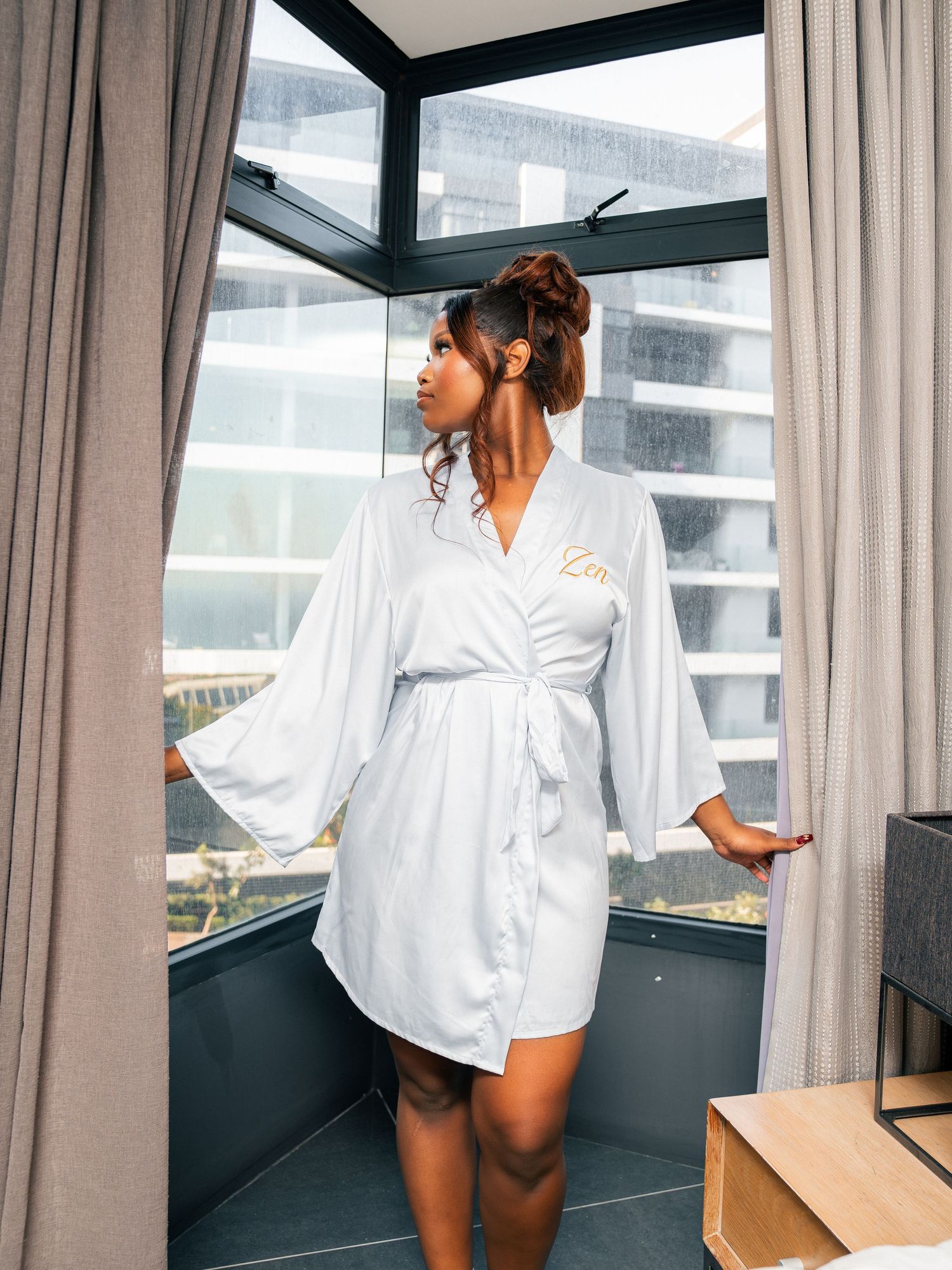 Woman in white personalized satin robe standing by large windows in a bright modern hotel room.