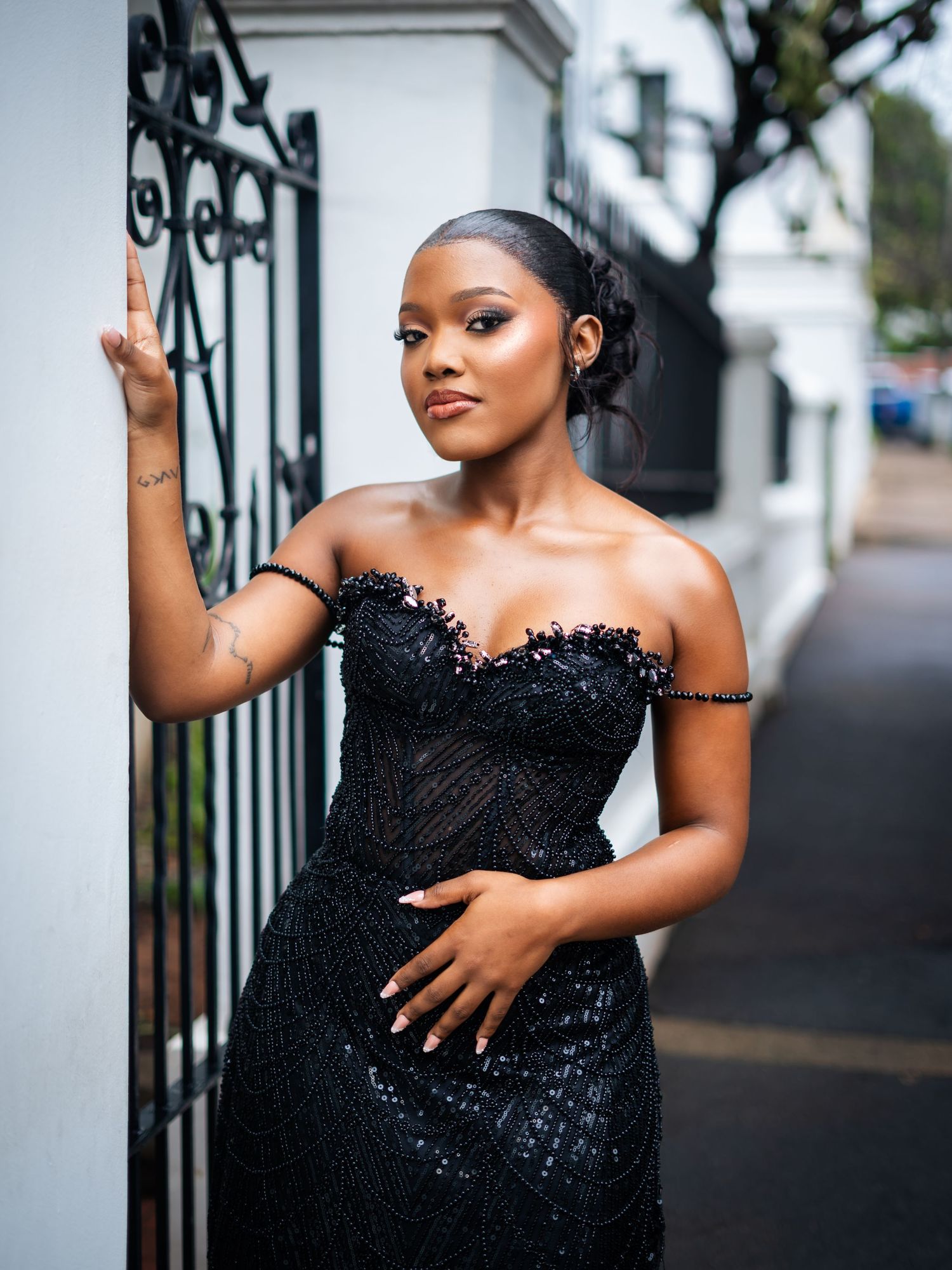 Woman in elegant black sequin off-shoulder gown posing gracefully by ornate iron gate and white wall.